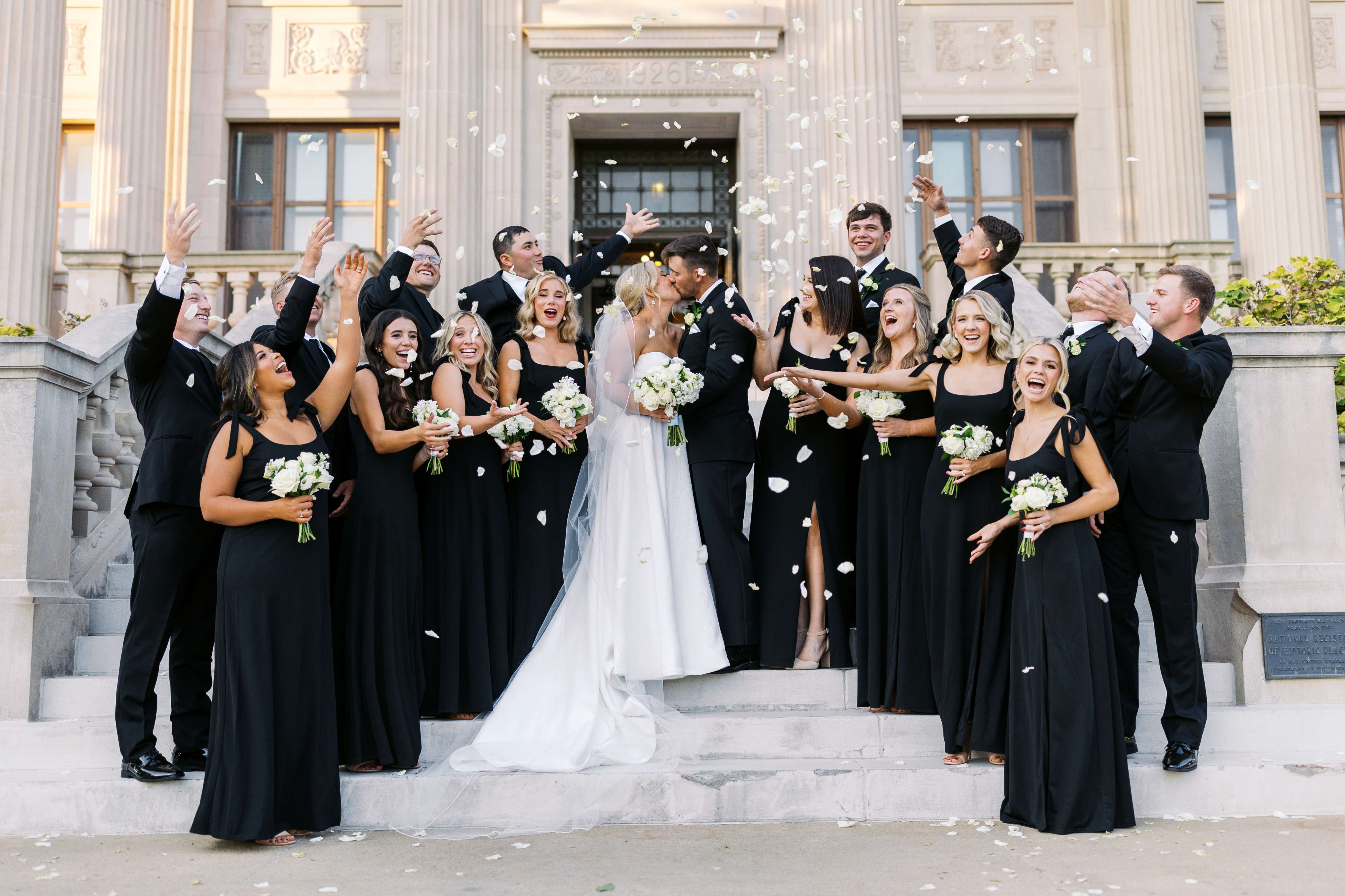 A bride and groom kiss on the front steps of the Oklahoma Hall of Fame, surrounded by the wedding party throwing rose petals.