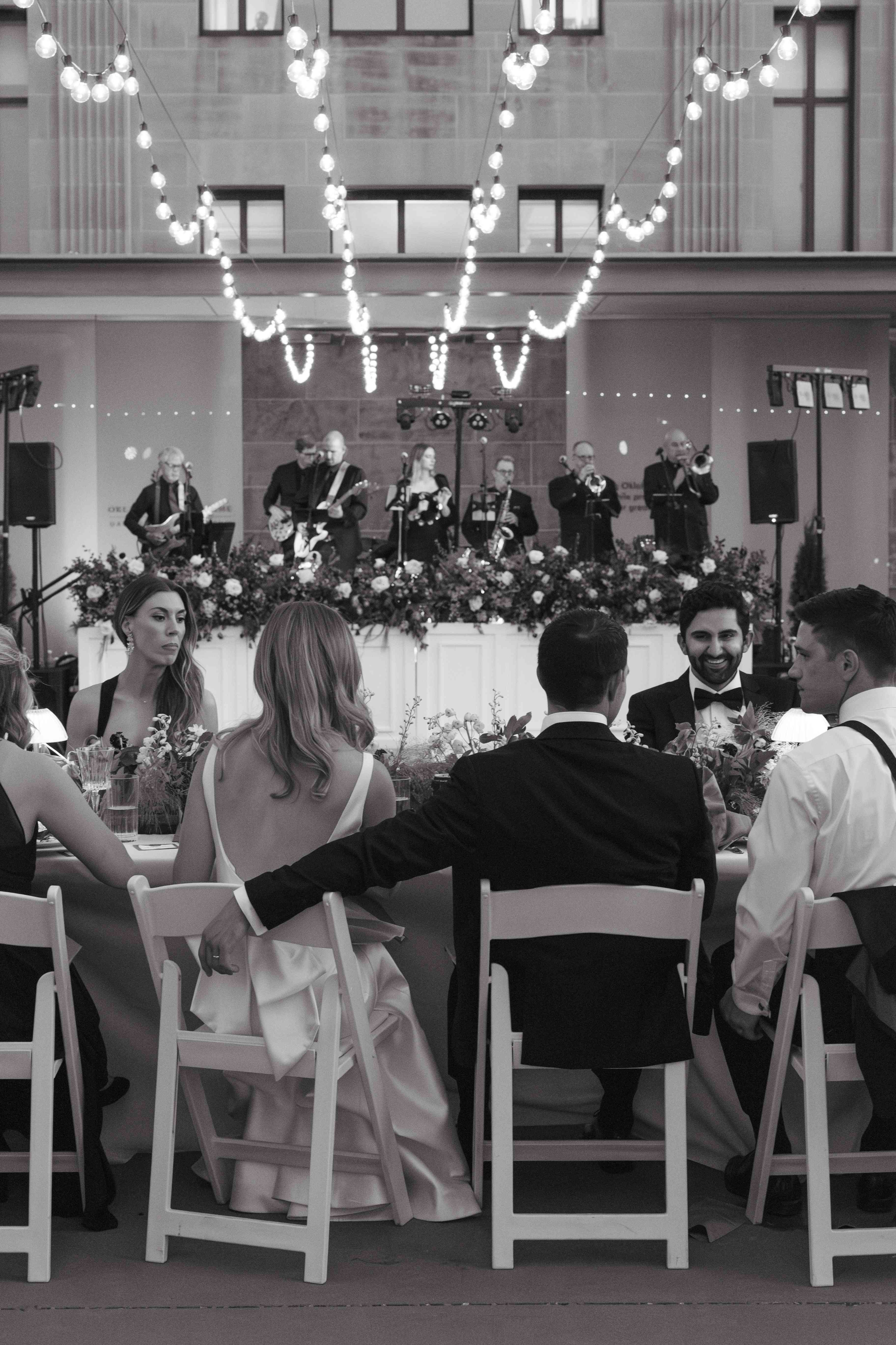 A bride and groom sit at a table in the Oklahoma Hall of Fame garden with musicians in the background.
