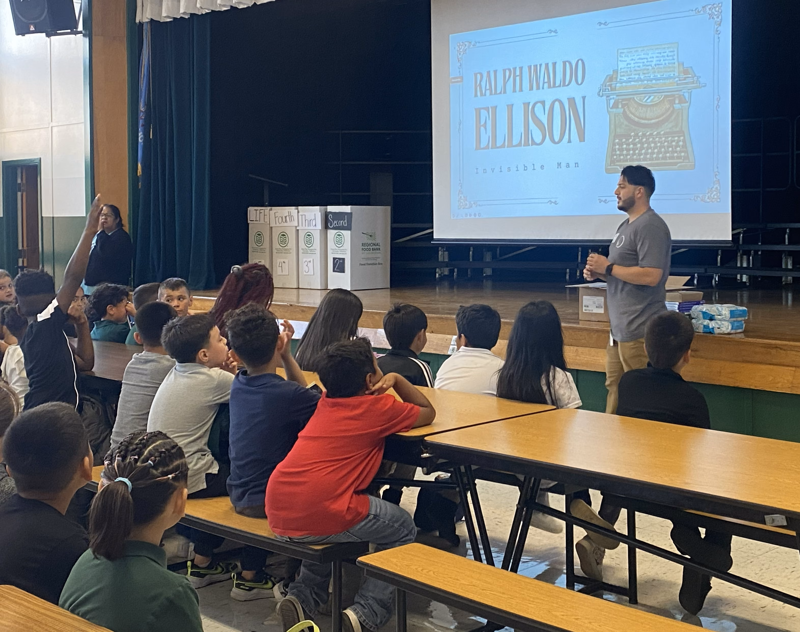 A Museum educator stands at the front of a school cafeteria, kicking off the Ralph Ellison Oklahoma Originals program. Students sit at lunch tables, listening.