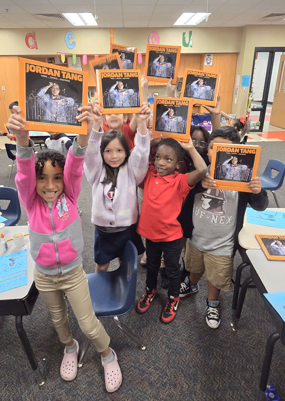 A group of elementary school children smile widely while each holding a Jordan Tang book above their heads.