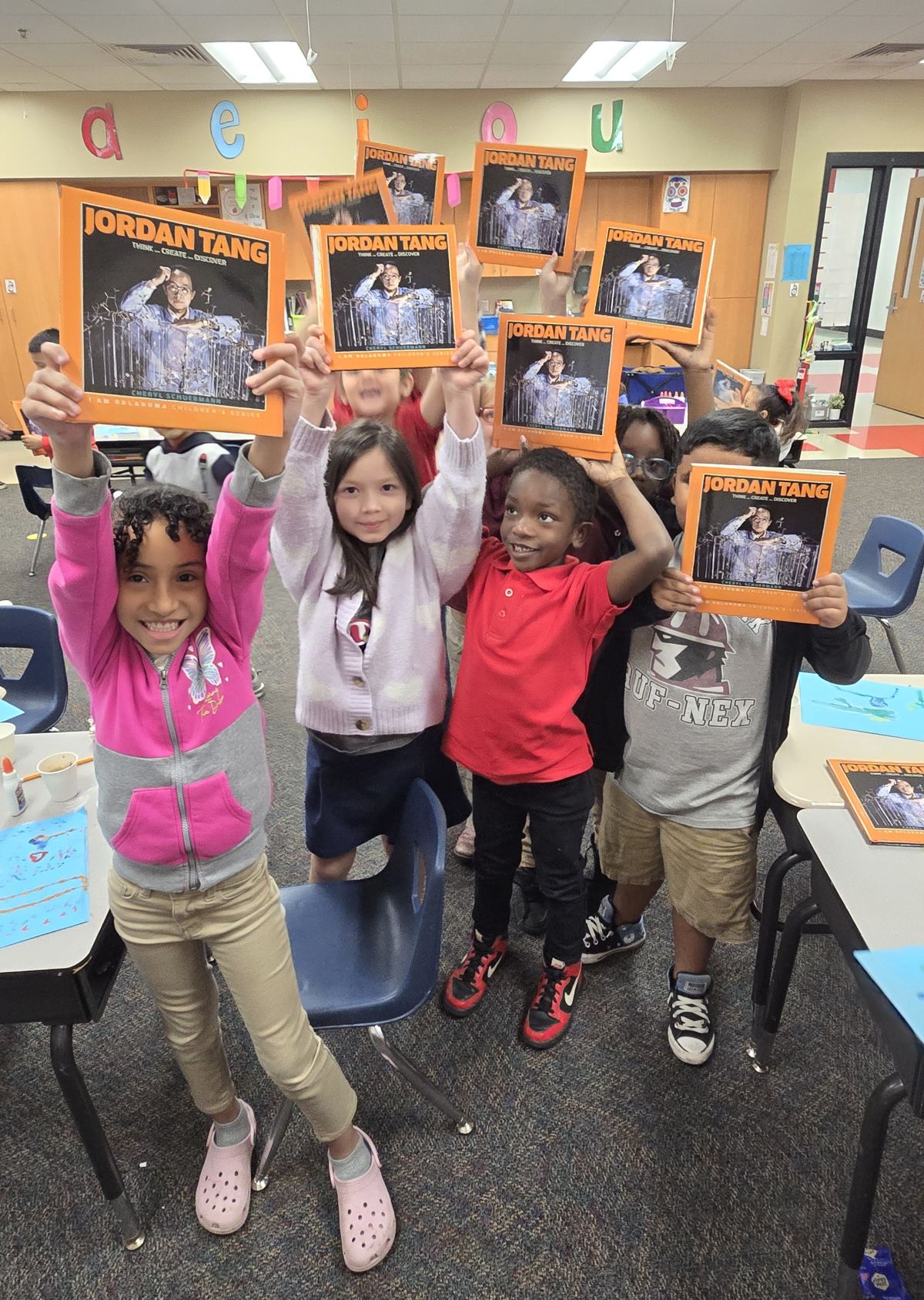 A group of elementary school children smile widely while each holding a Jordan Tang book above their heads.