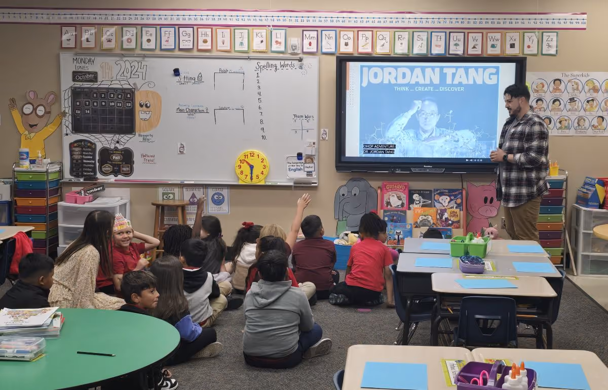 A Museum educator stands at the front of a classroom, talking to a group of elementary school students, seated on the floor. An image of Jordan Tang is displayed on the screen behind him.