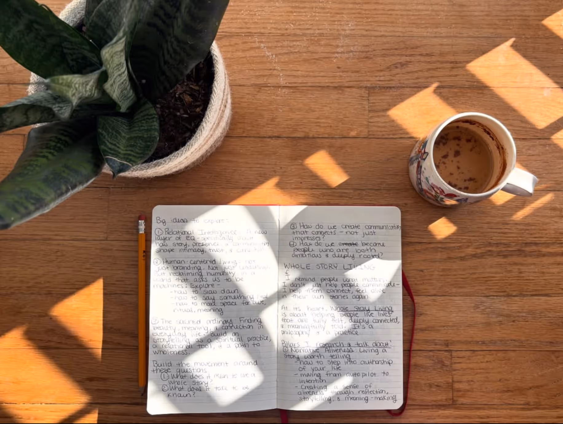 Open notebook with handwritten notes, pencil on its left, potted plant on top left, and coffee cup on right on a wooden floor with sunlight shadows.