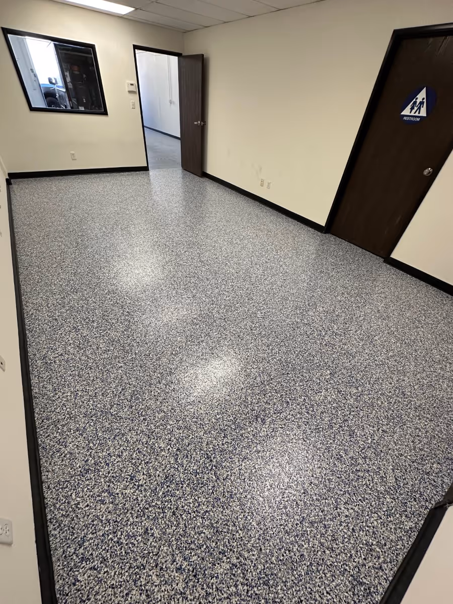 Empty room with speckled epoxy flooring, beige walls, dark wooden doors, and a window showing an outdoor truck.