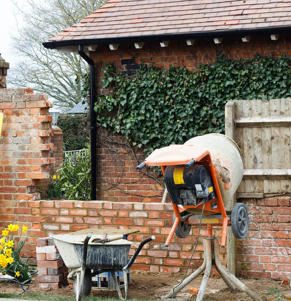 Cement mixer, pug board and wheelbarrow by wall and out house.
