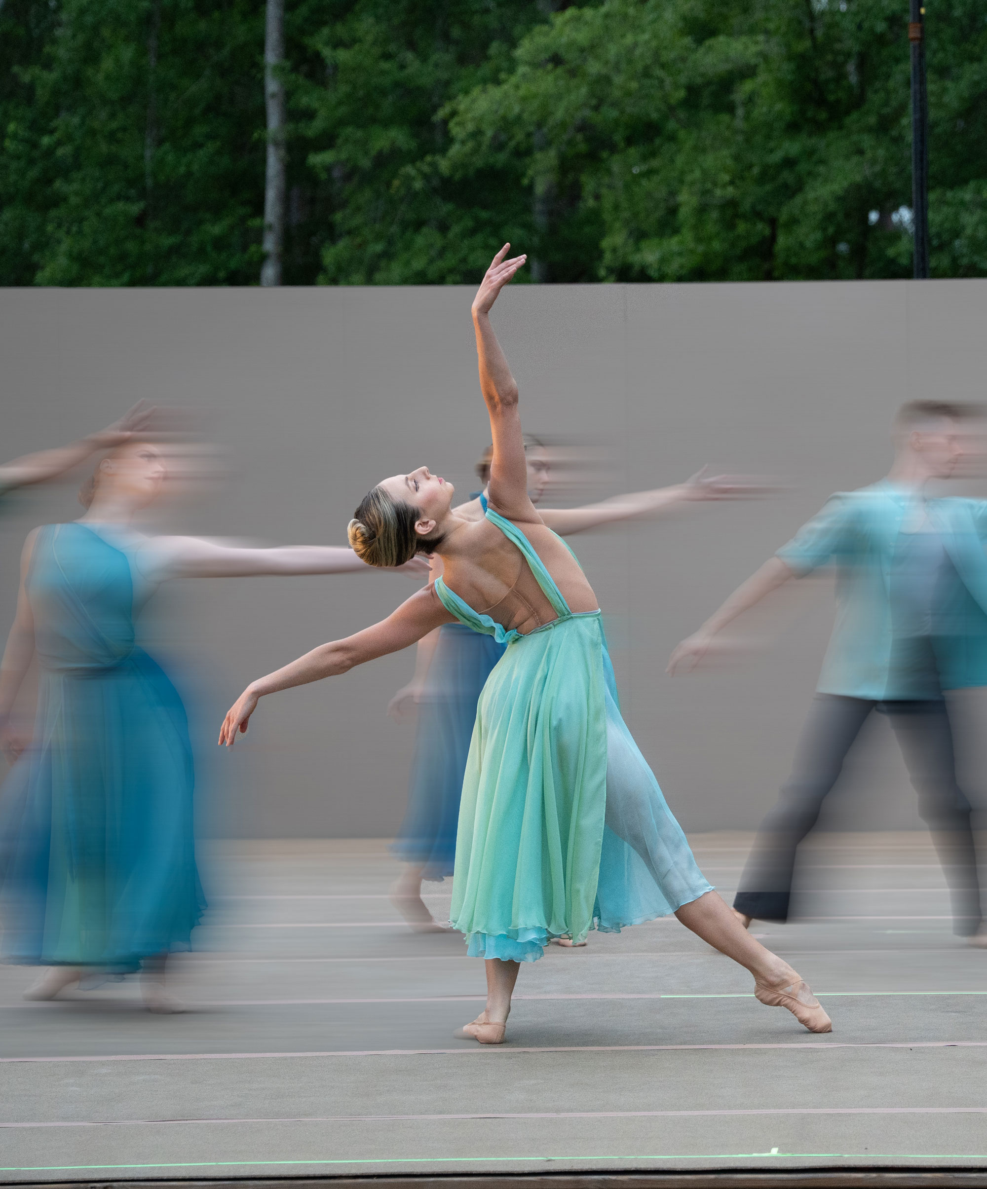 Ballet dancer in flowing aqua dress performing outdoors with motion-blurred ensemble