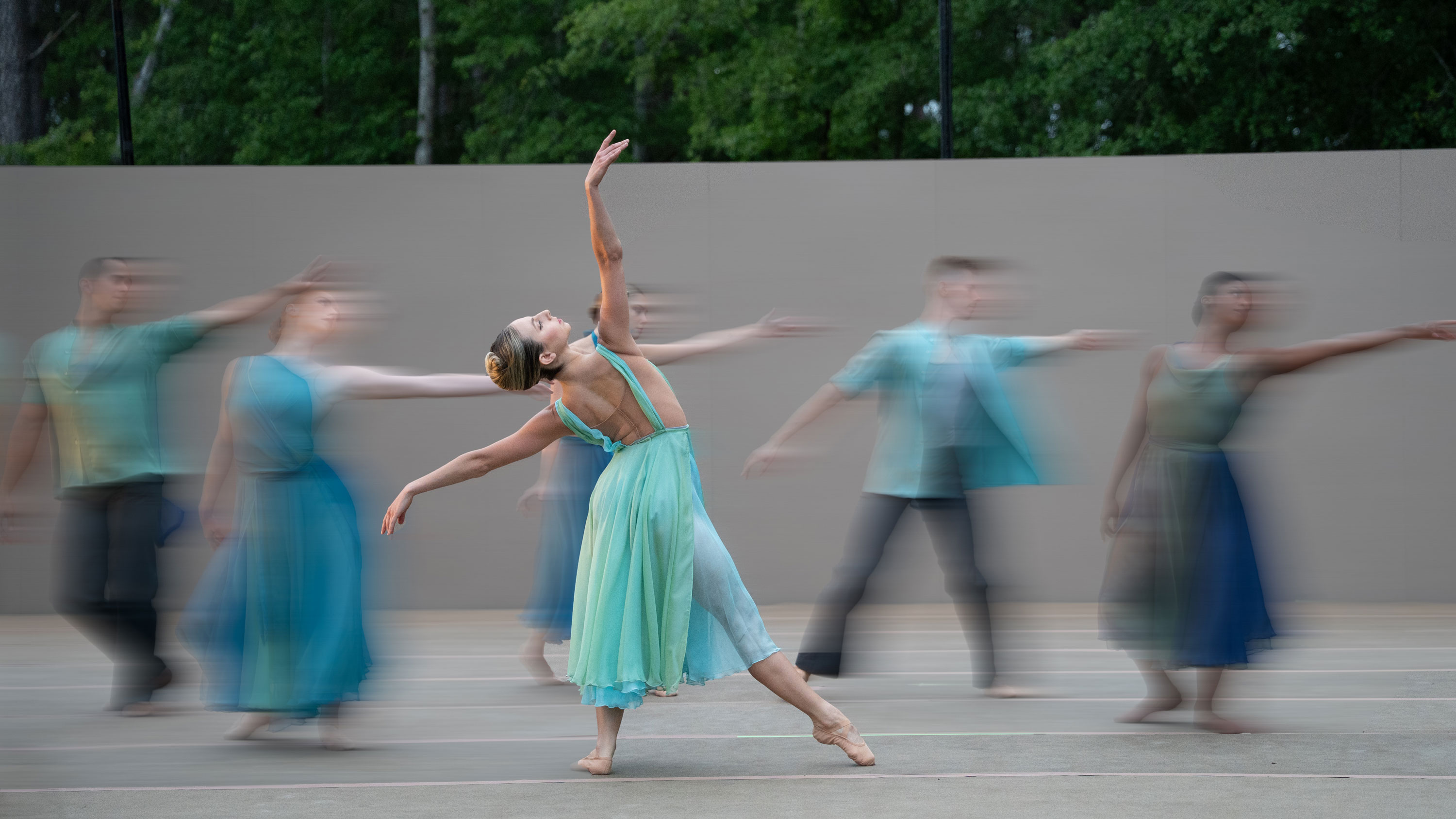 Ballet dancer in flowing aqua dress performing outdoors with motion-blurred ensemble