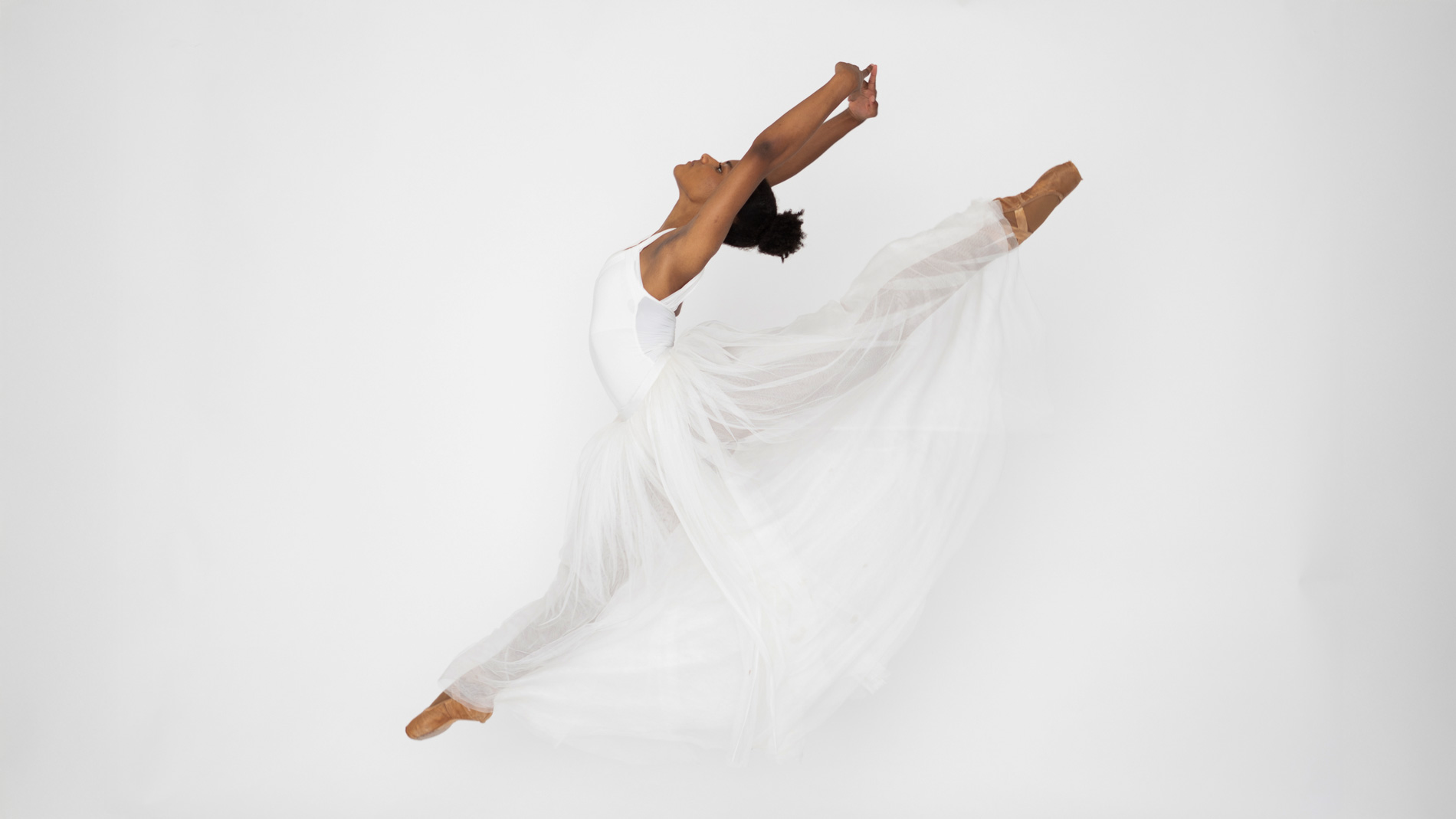 Black ballerina in white leotard and tulle skirt performing a grand jeté in studio