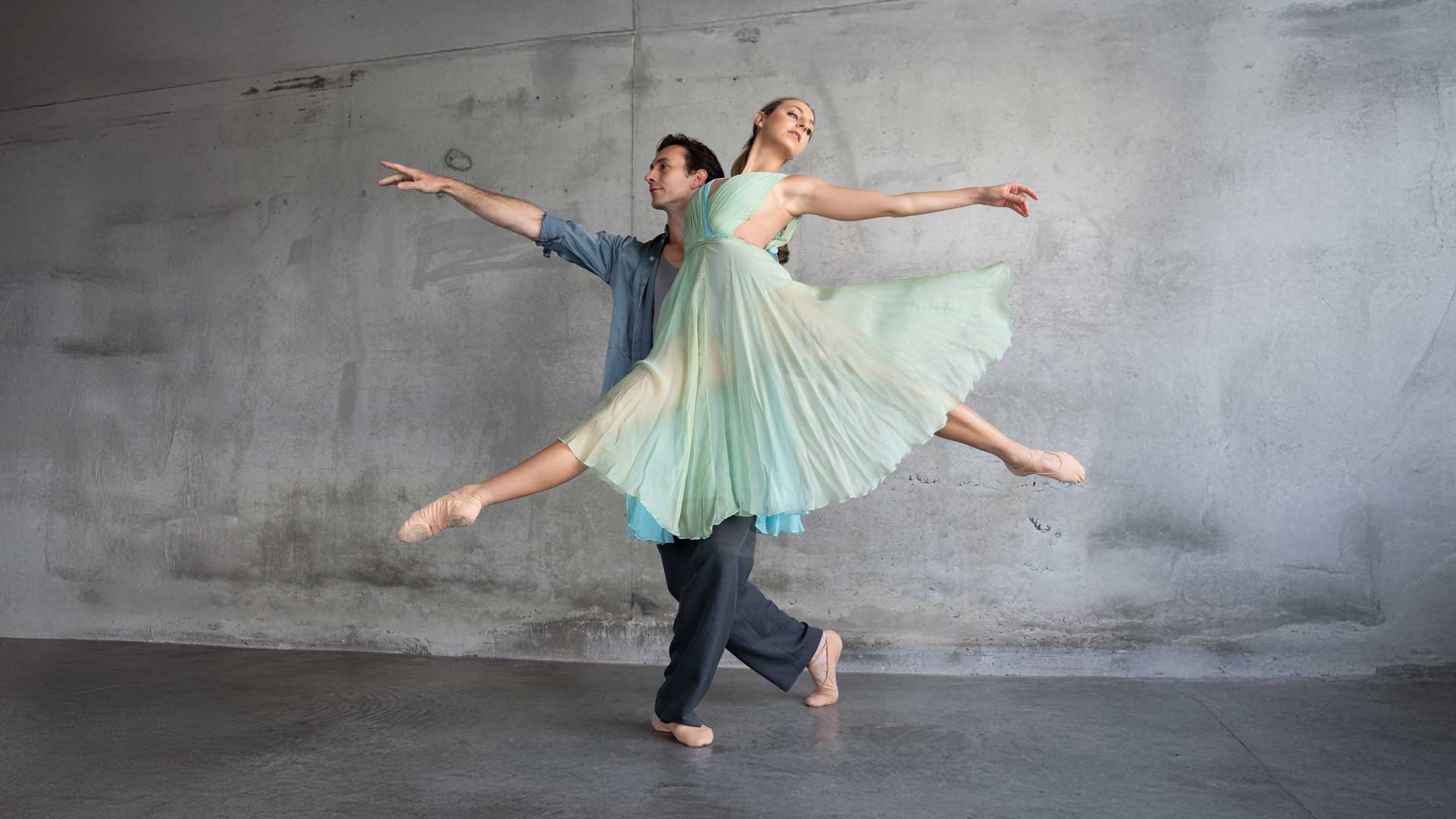 Two ballet dancers performing a graceful lift in a studio with concrete walls.