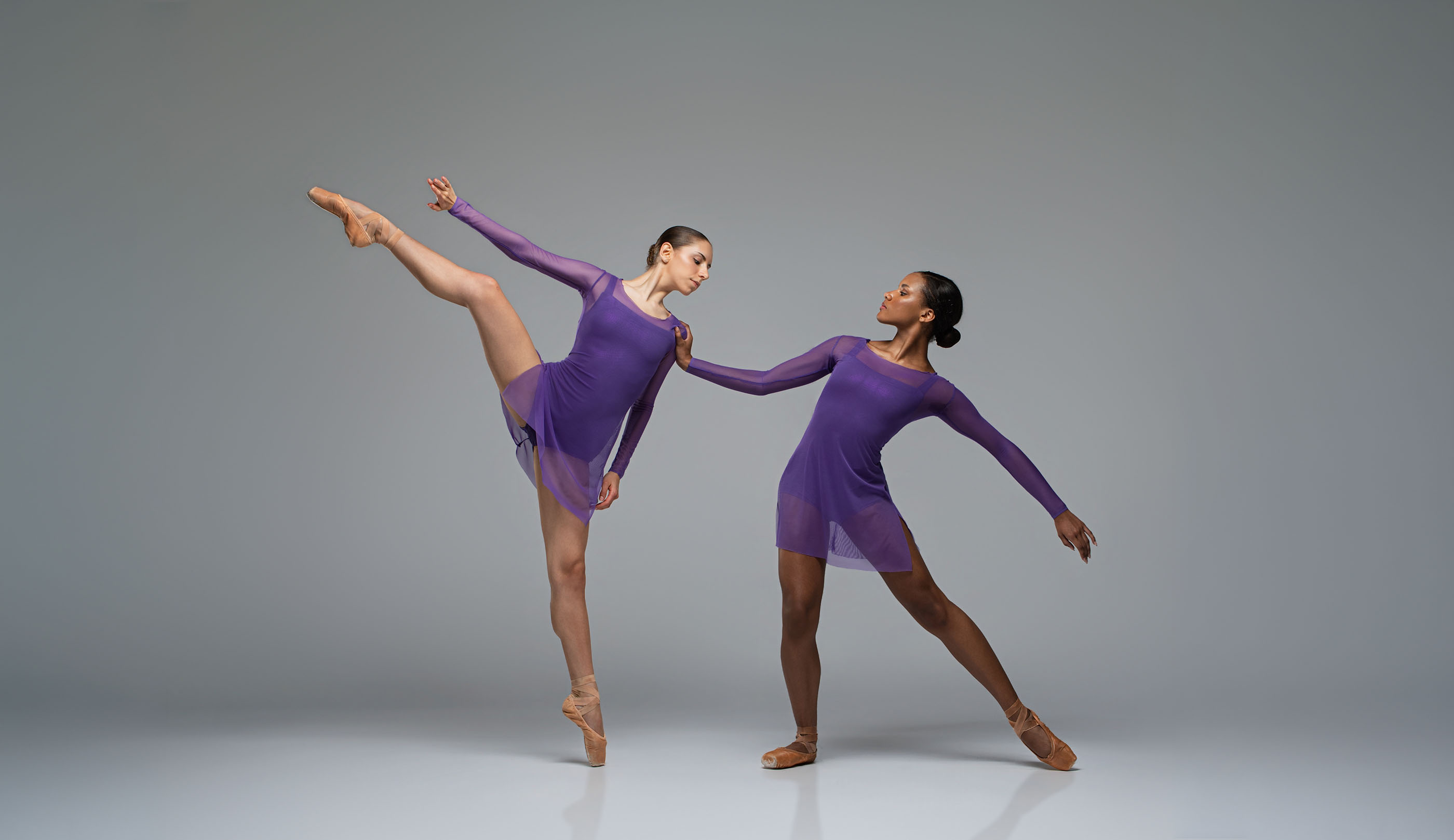 Two ballet dancers in sheer purple dresses balance en pointe with one leg extended horizontally and one arm raised, posing symmetrically against a plain gray studio background.