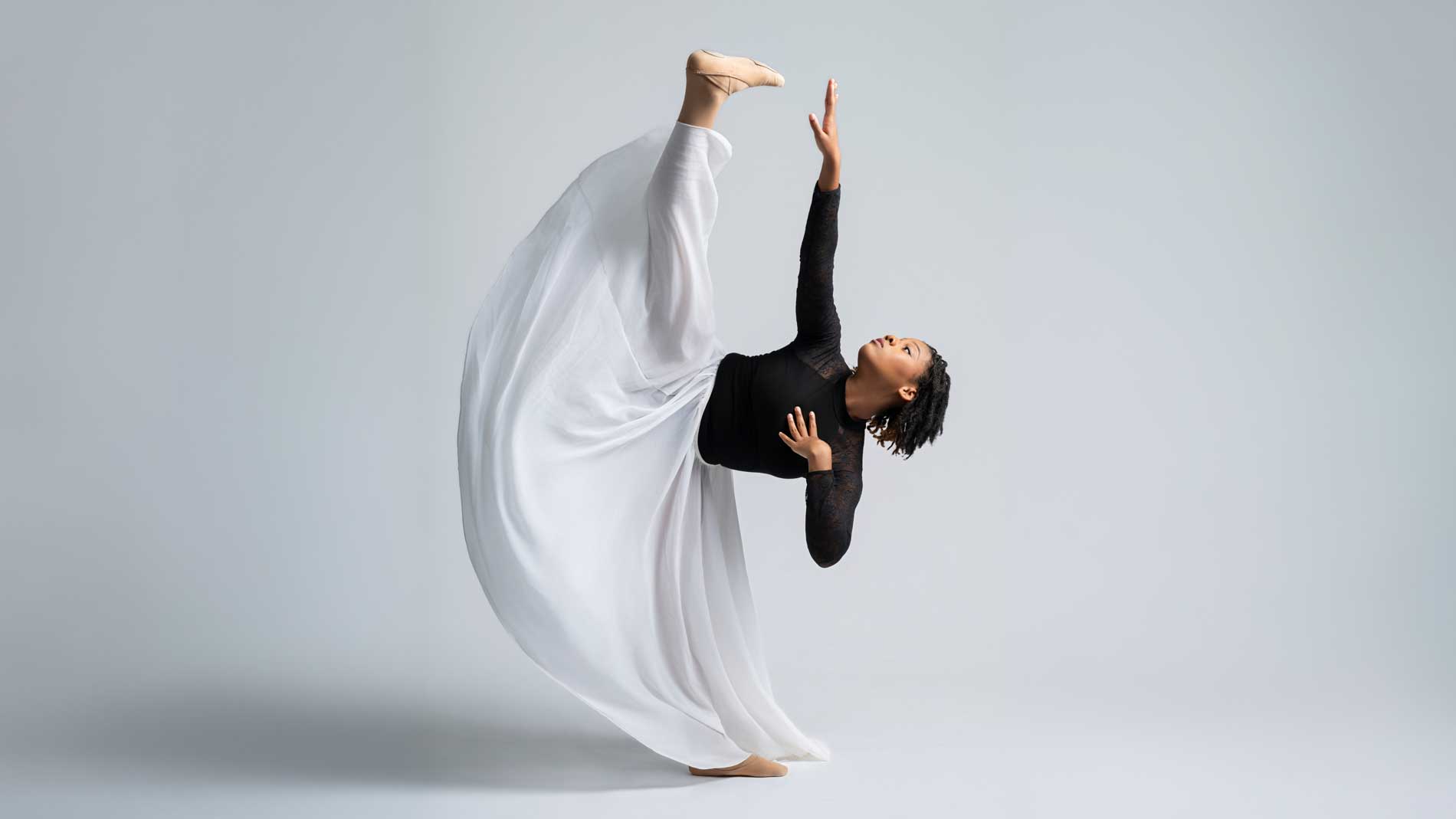Dancer in a black top and flowing white skirt performing a high leg extension.