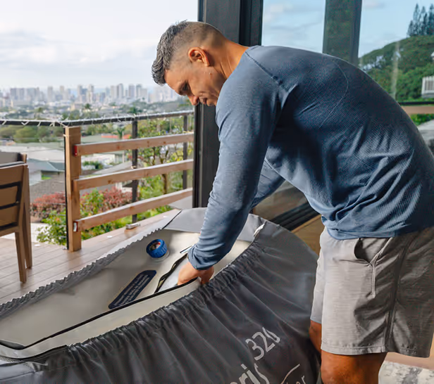 Man in blue long-sleeve shirt and shorts preparing a hyperbaric chamber indoors near a glass door with an outdoor deck and city view.