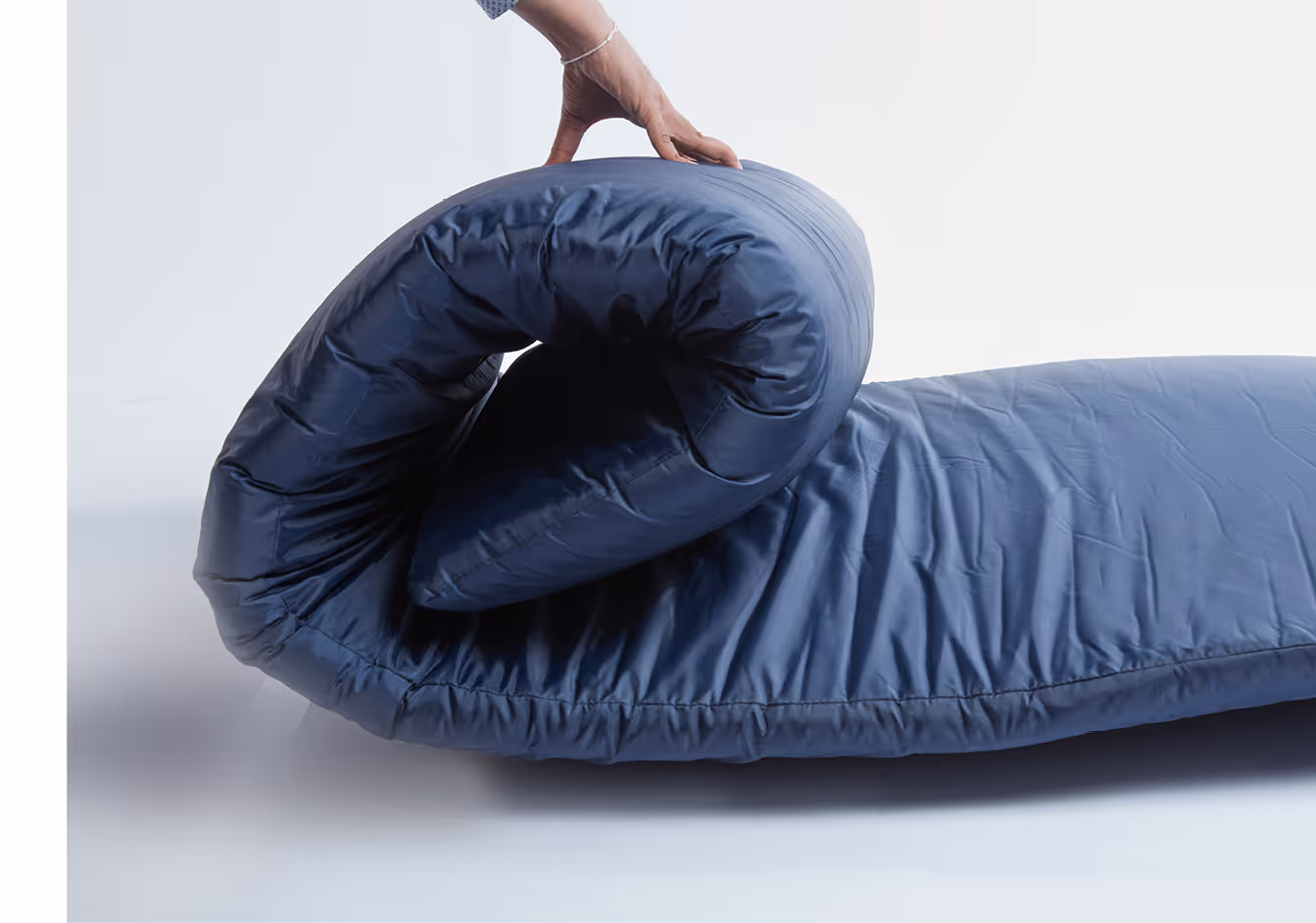 Hand rolling up a navy blue sleeping bag on a white surface.