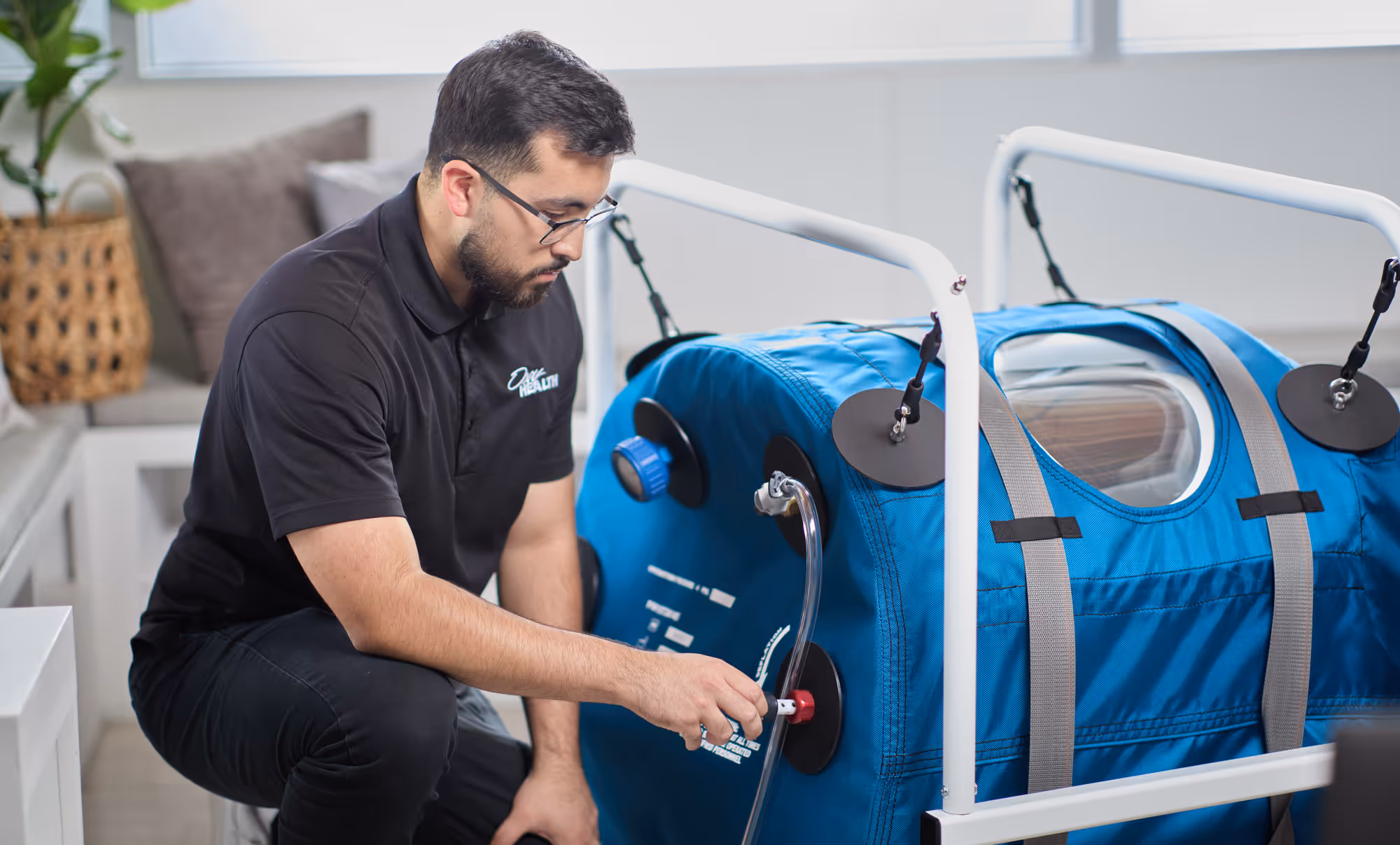 Man wearing glasses adjusting a valve on a blue medical hyperbaric oxygen chamber in a bright room.