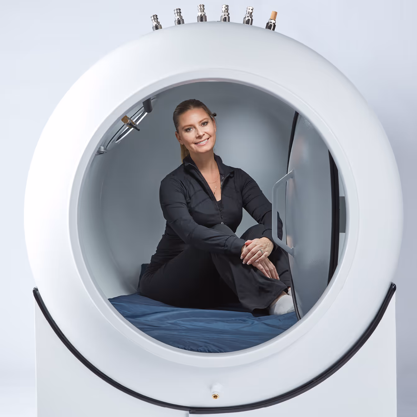 Woman dressed in black sitting inside a white hyperbaric oxygen chamber with a blue mattress.