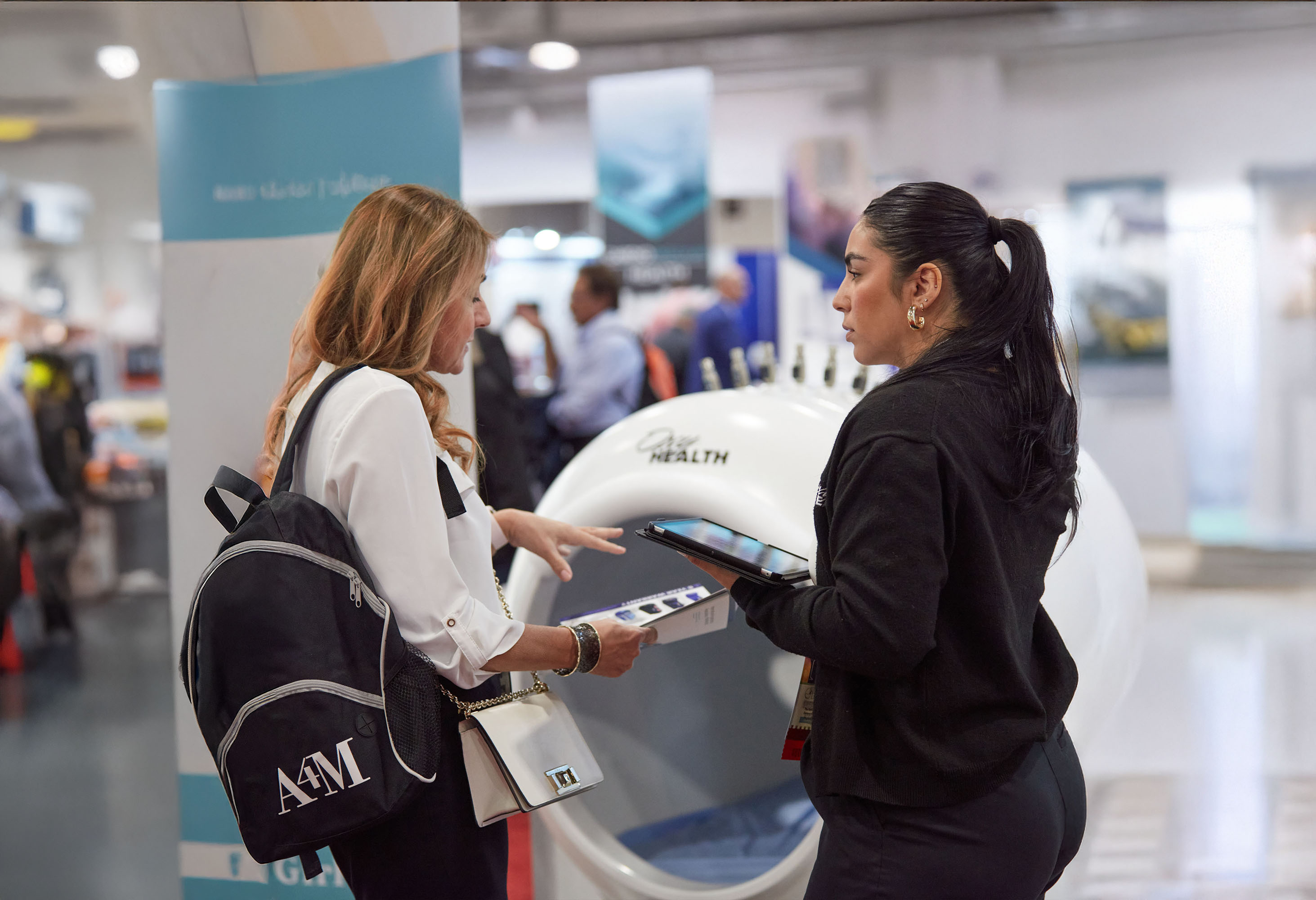 Two women engaged in conversation at A4m event, one holding a tablet and the other holding a brochure, with exhibition displays in the background.