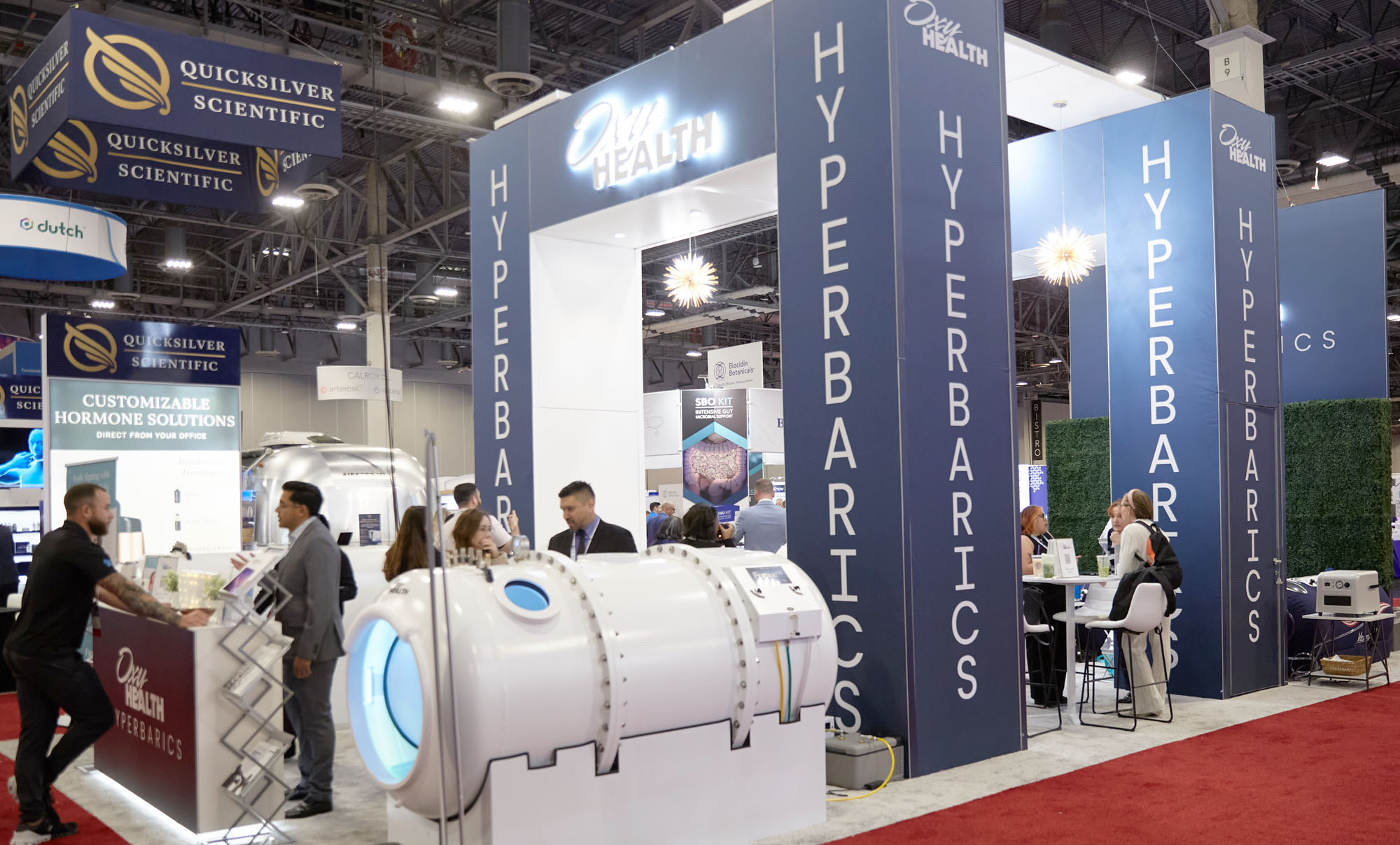 Trade show booth for Oxy Health Hyperbarics featuring a large white hyperbaric chamber and people interacting at the display.