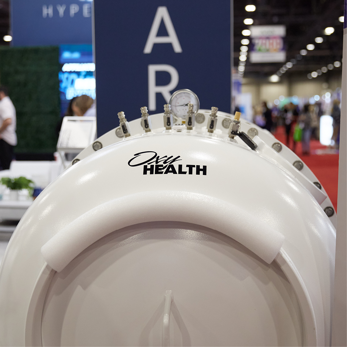 Close-up of a Fortius 420 hyperbaric oxygen chamber with pressure gauges and valves labeled 'Oxy Health' at a trade show.