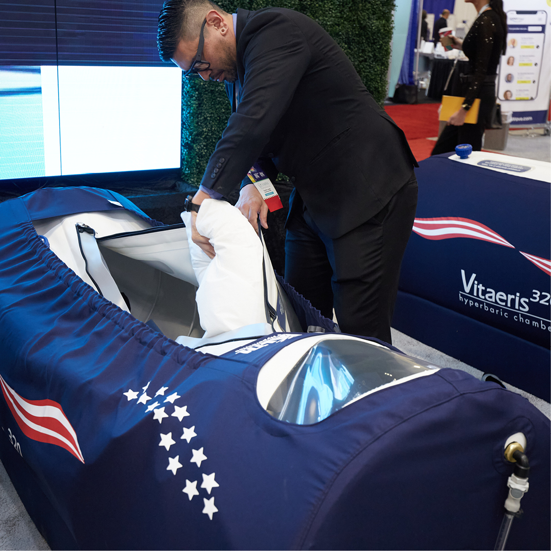 Man in a black suit adjusting the interior padding of a navy blue hyperbaric chamber labeled Vitaeris 320 at an exhibition.