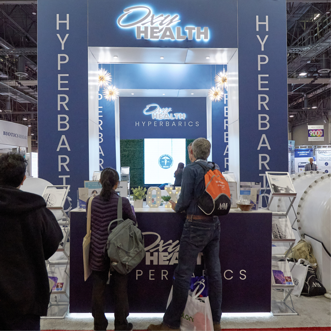 Trade show booth for Oxy Health Hyperbarics with three people standing in front and promotional materials displayed.