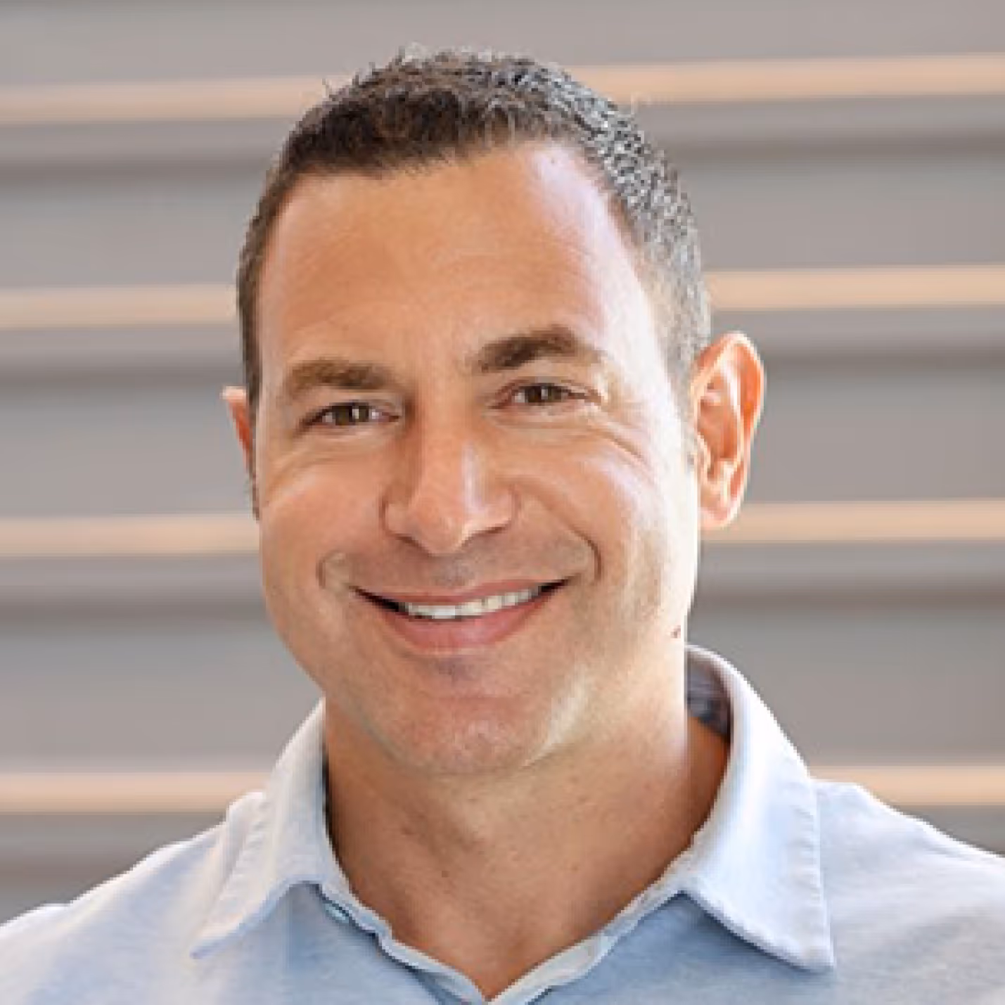 Dr. Sonners smiling with short dark hair wearing a light blue collared shirt in front of gray stairs background.
