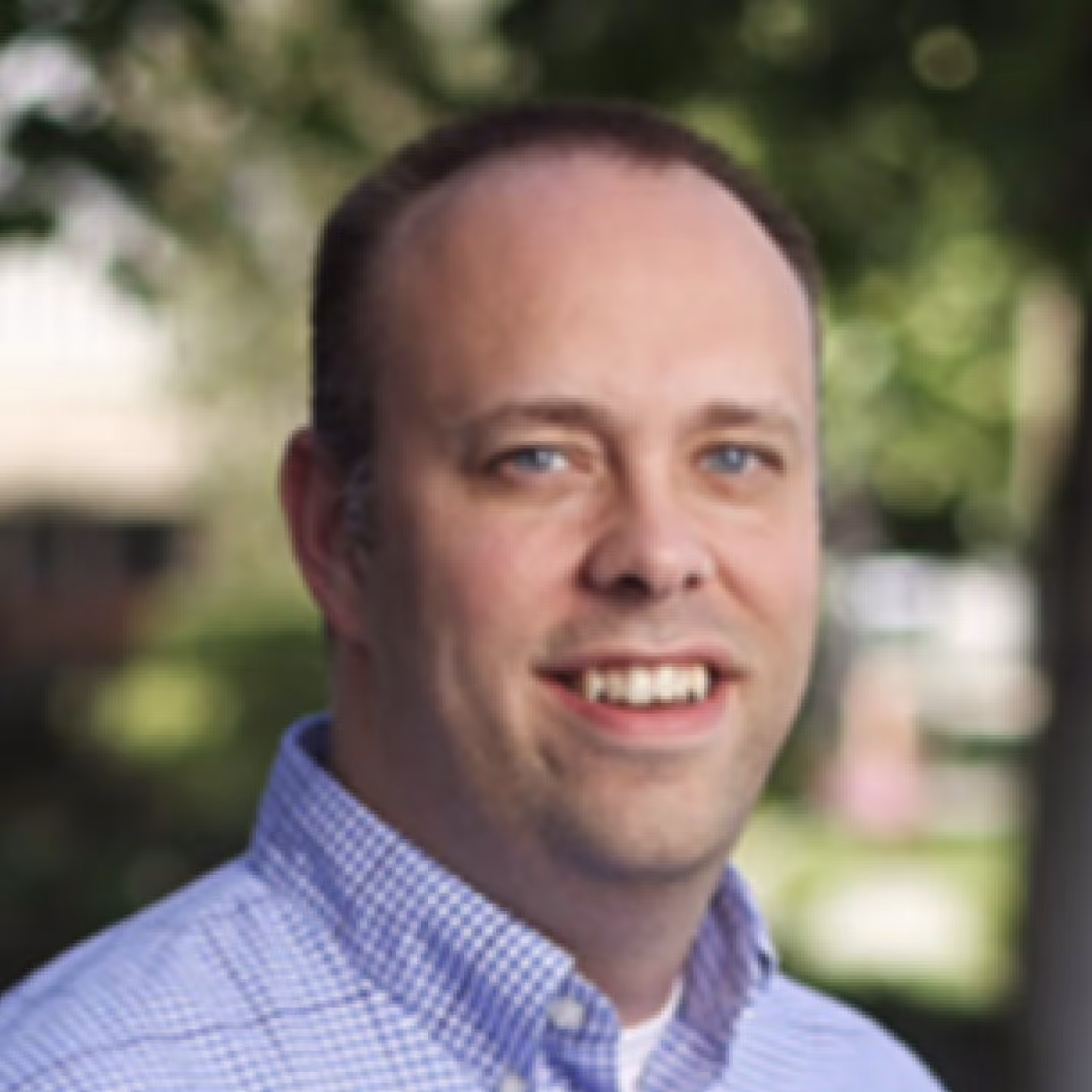 Dr. Rossignol with blue eyes wearing a blue checkered shirt outdoors with greenery background.