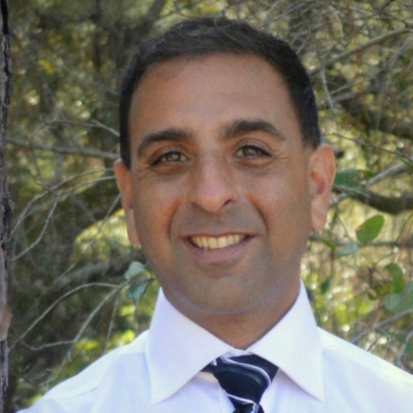 Dr. Ratansi in white shirt and striped tie with trees in the background.
