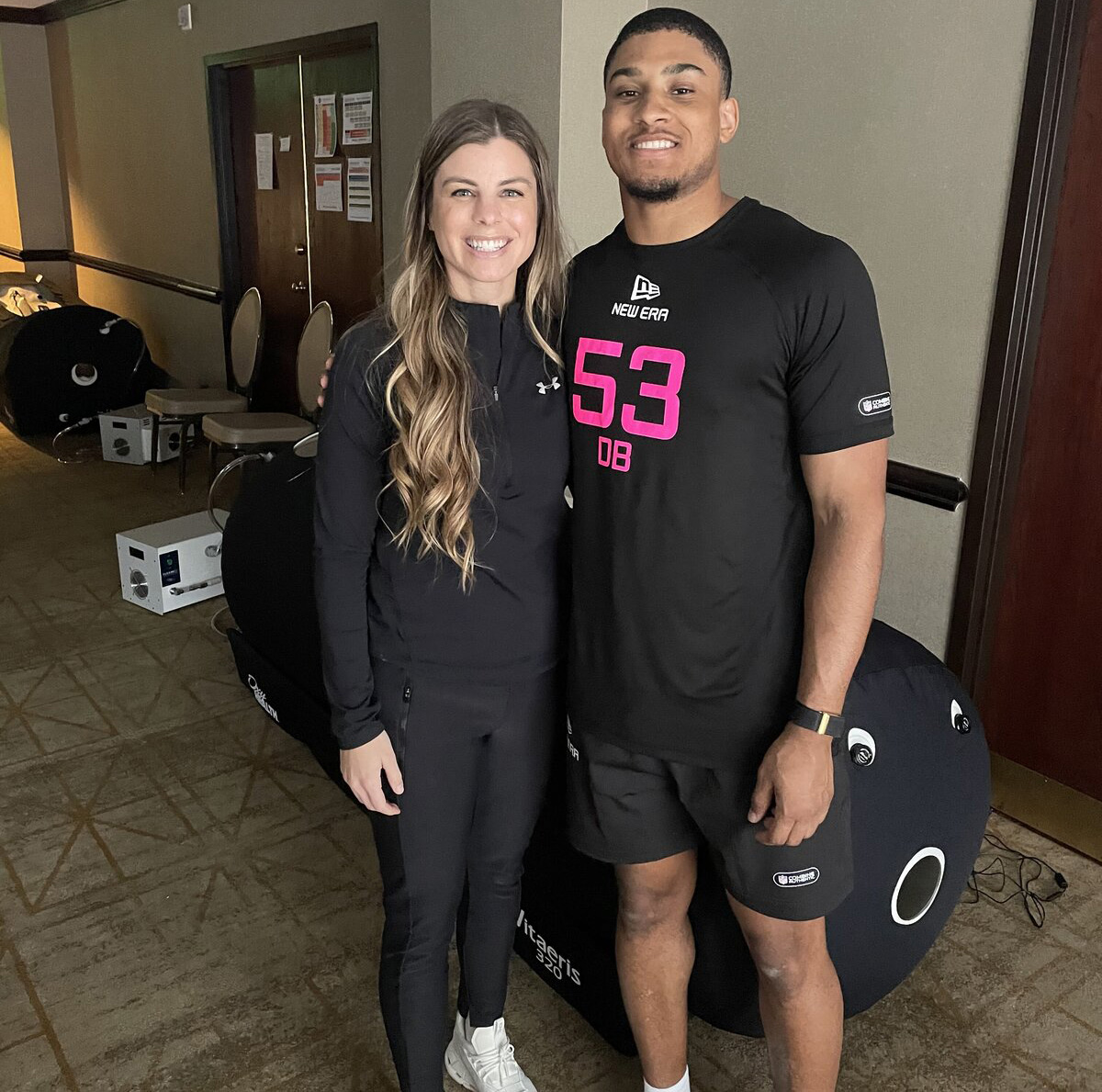 Dr. Tracy with long wavy hair in black athletic wear standing next to smiling man in black sportswear with number 53 on his shirt indoors.