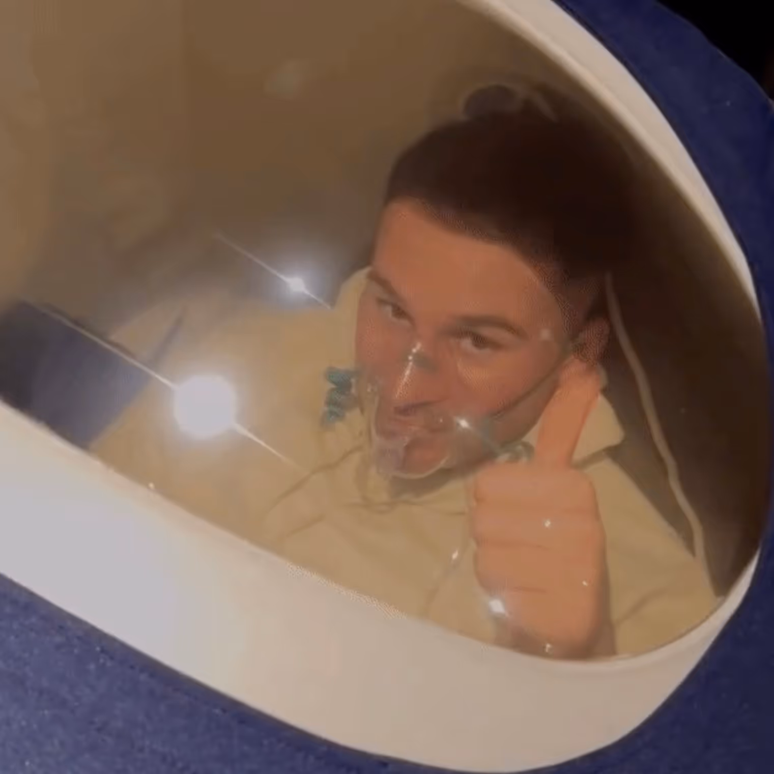Alexis mac allister inside a Vitaeris 320 hyperbaric oxygen chamber wearing an oxygen mask and giving a thumbs-up.