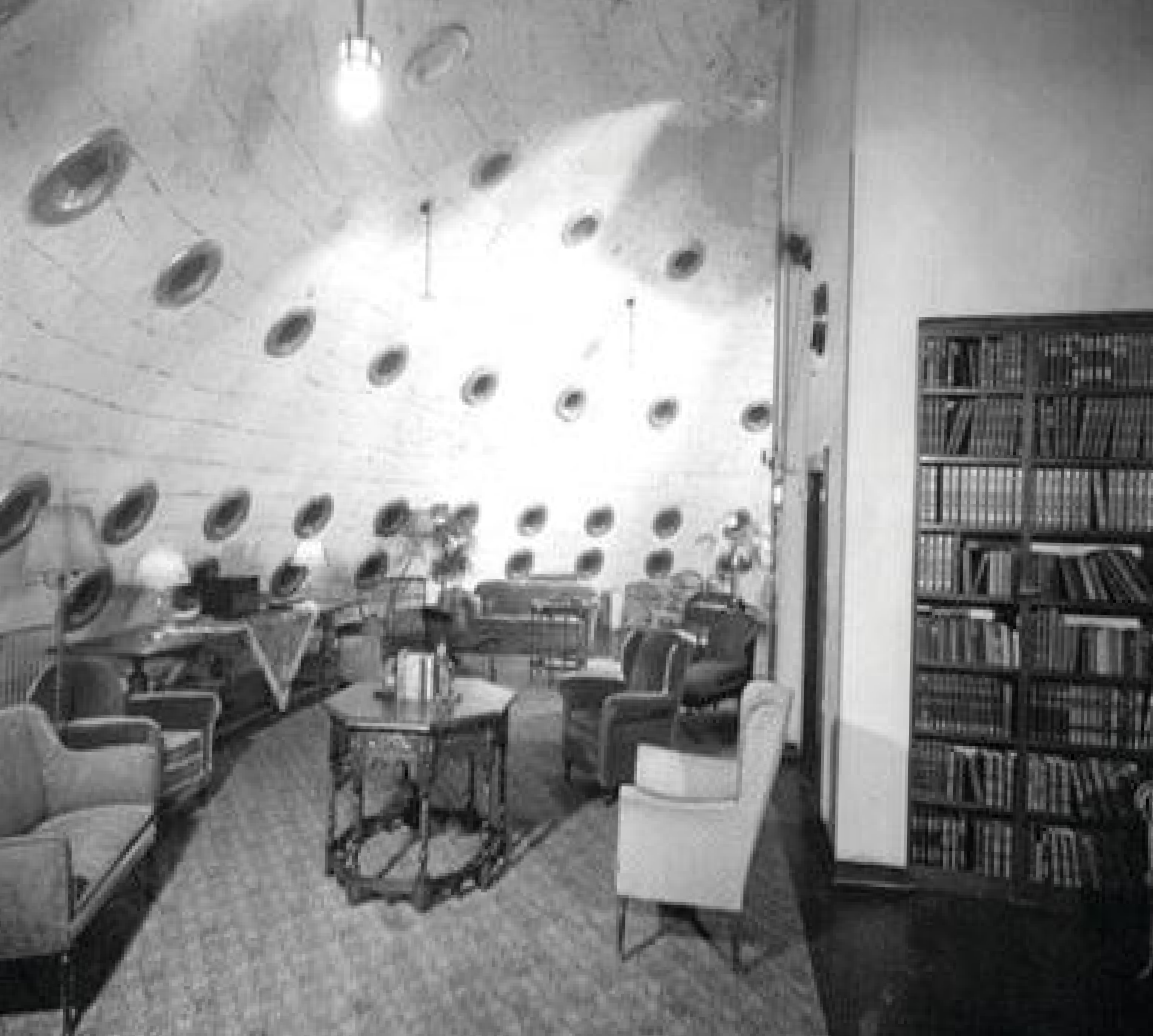 Black and white image of a circular hyperbaric chamber room with round windows, furnished with armchairs, couches, tables, lamps, and bookshelves.