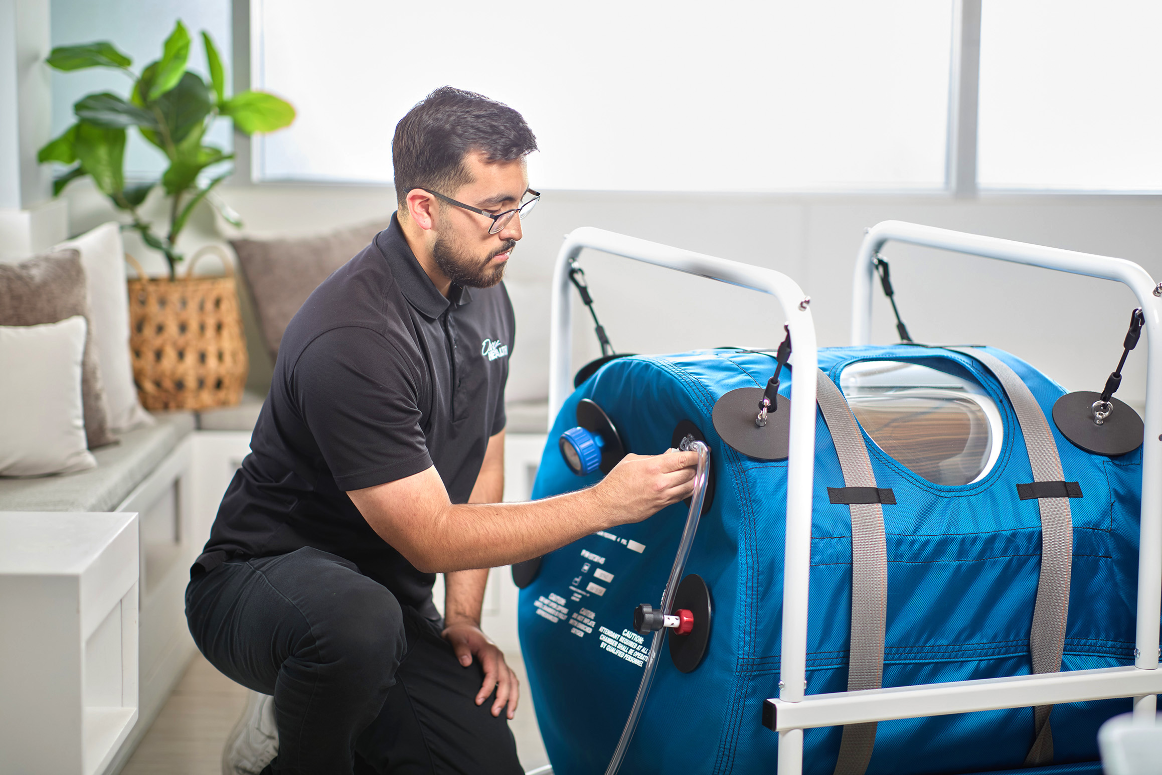 Man in black shirt with glasses kneeling and adjusting a Quamvis 320 hyperbaric oxygen chamber inside a bright room.