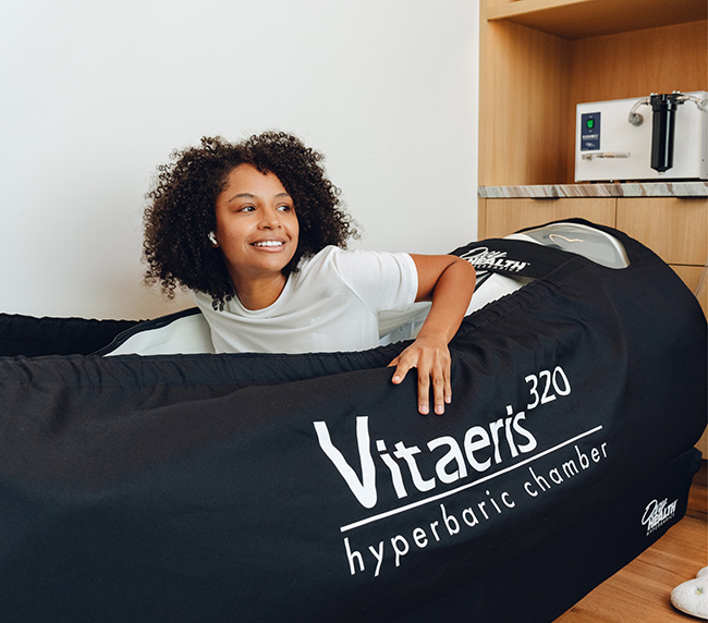 Smiling woman sitting inside a Vitaeris 320 portable hyperbaric chamber in a room with wooden cabinets.