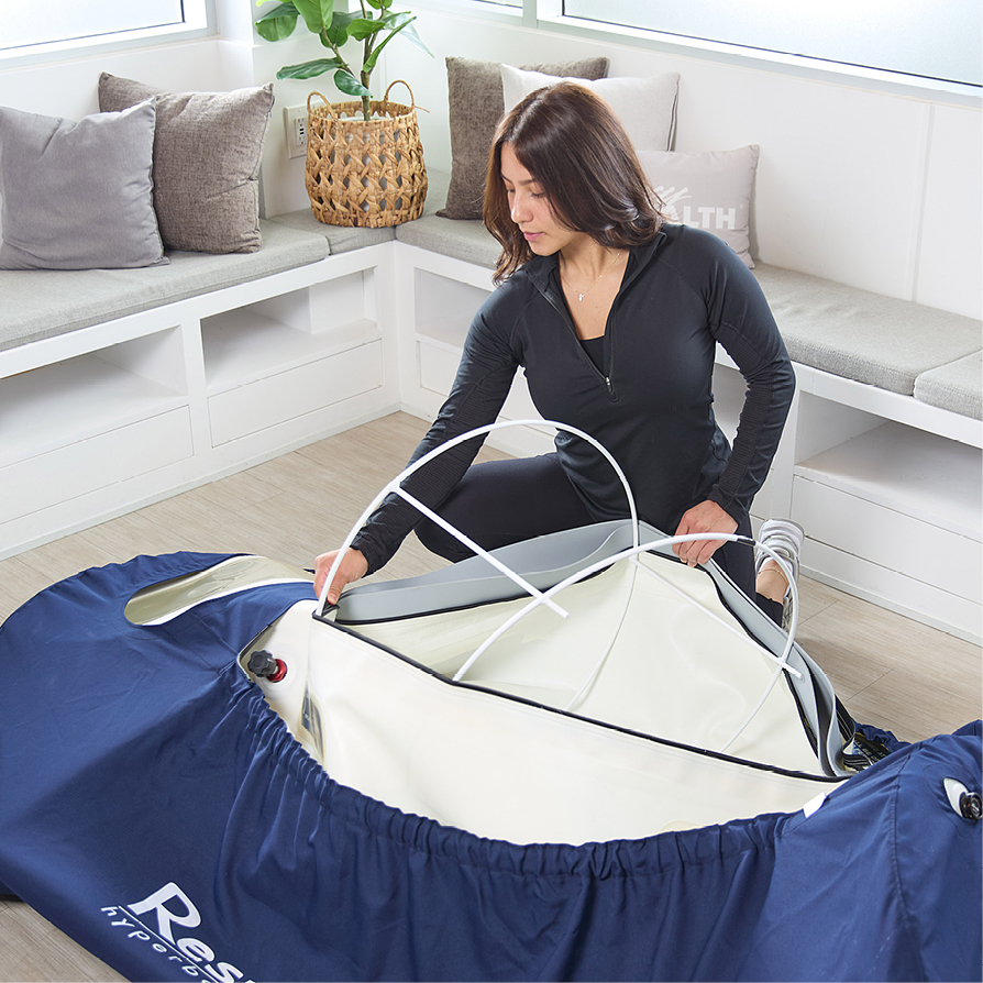 Woman in black athletic wear assembling or adjusting a large blue and white Respiro hyperbaric chamber in a bright room with cushioned bench seating and pillows.
