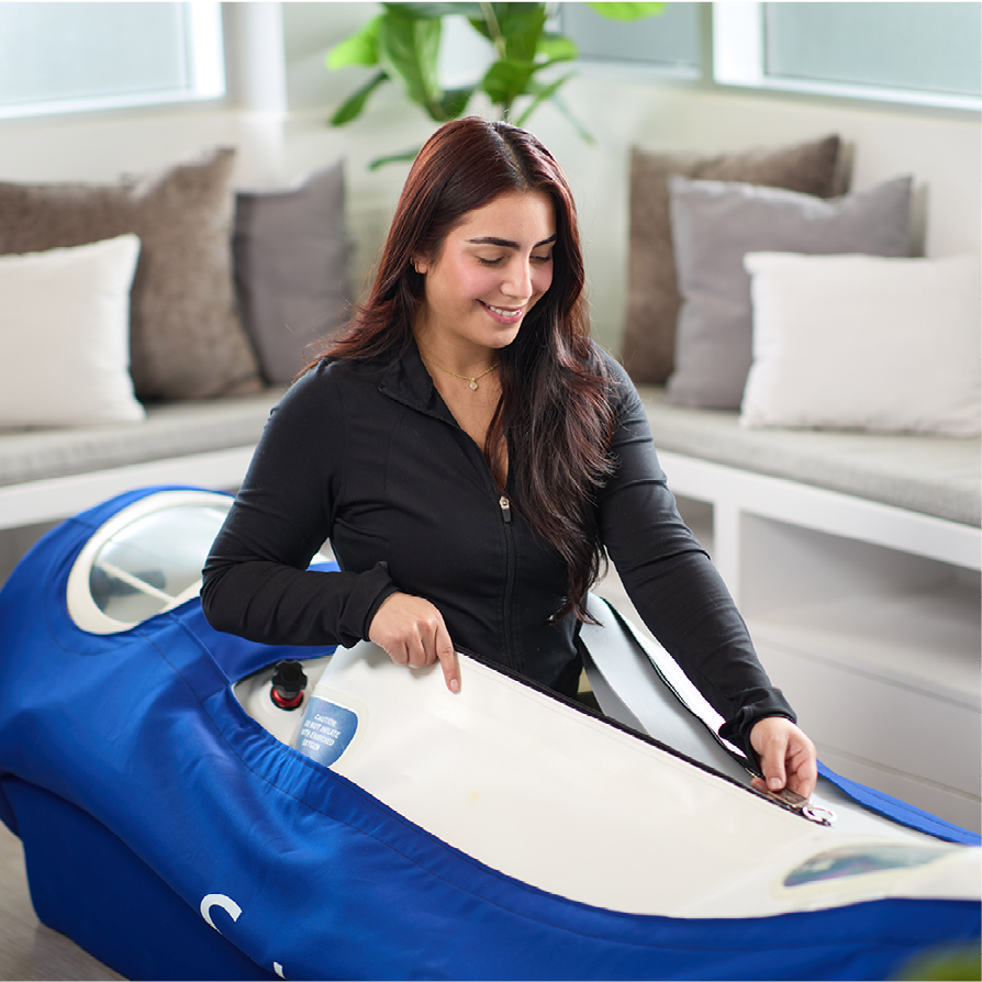 Smiling woman in a black jacket unzips a blue hyperbaric oxygen therapy chamber in a bright, modern room.