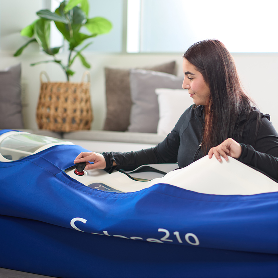 Woman using a blue and white Thermae 210 sauna blanket in a cozy room with a plant and cushions in the background.