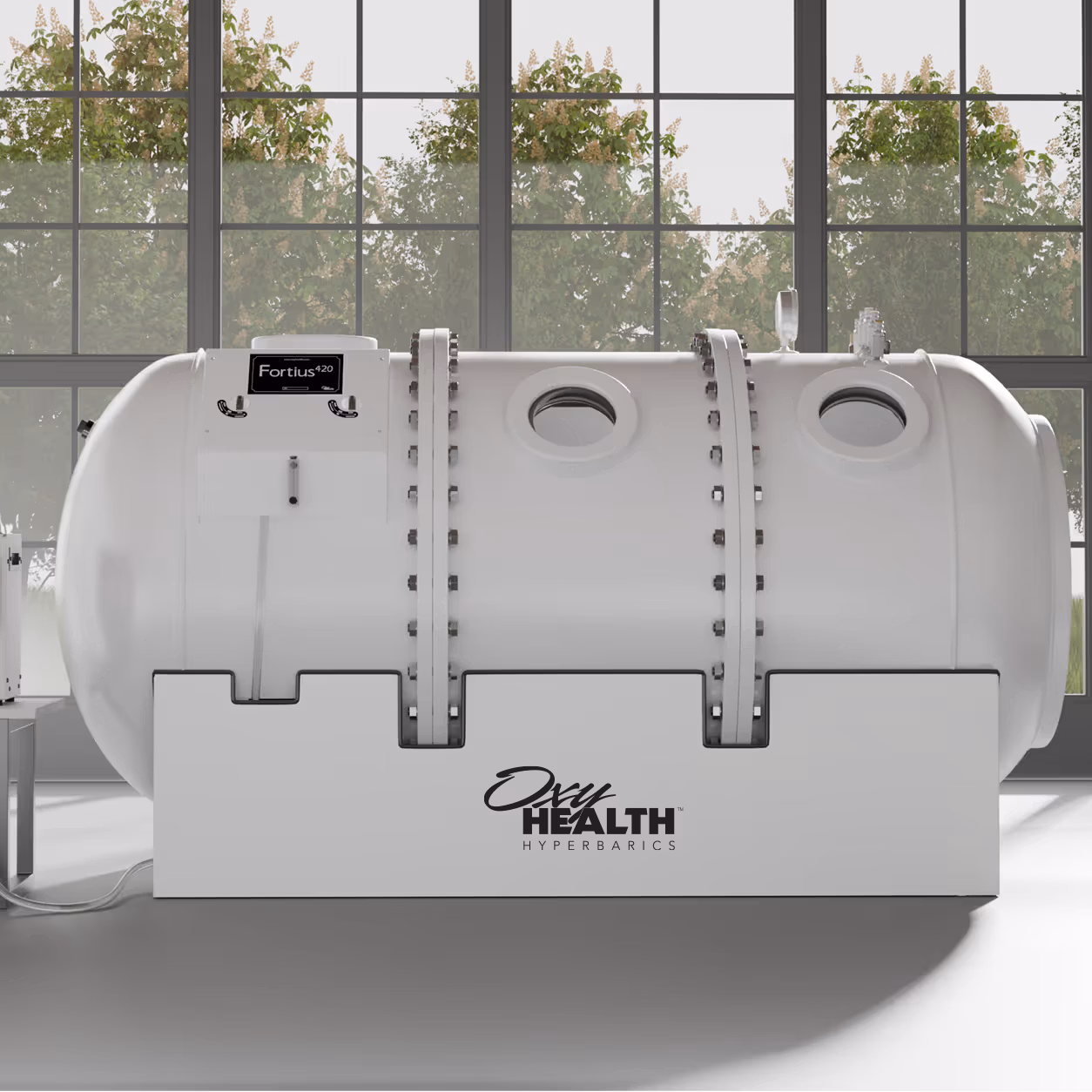 White cylindrical Fortius 420 hyperbaric chamber with two round windows in a bright room with large windows and green trees outside.