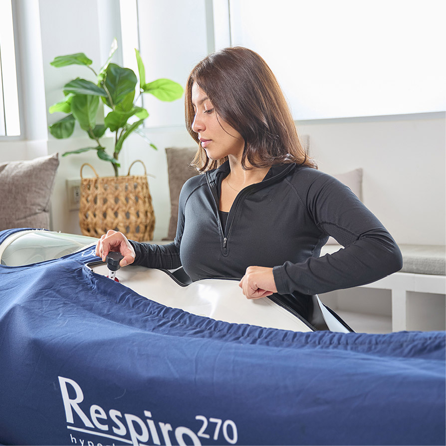 Woman adjusting a Respiro270 hyperbaric chamber in a bright room with plants and cushions.