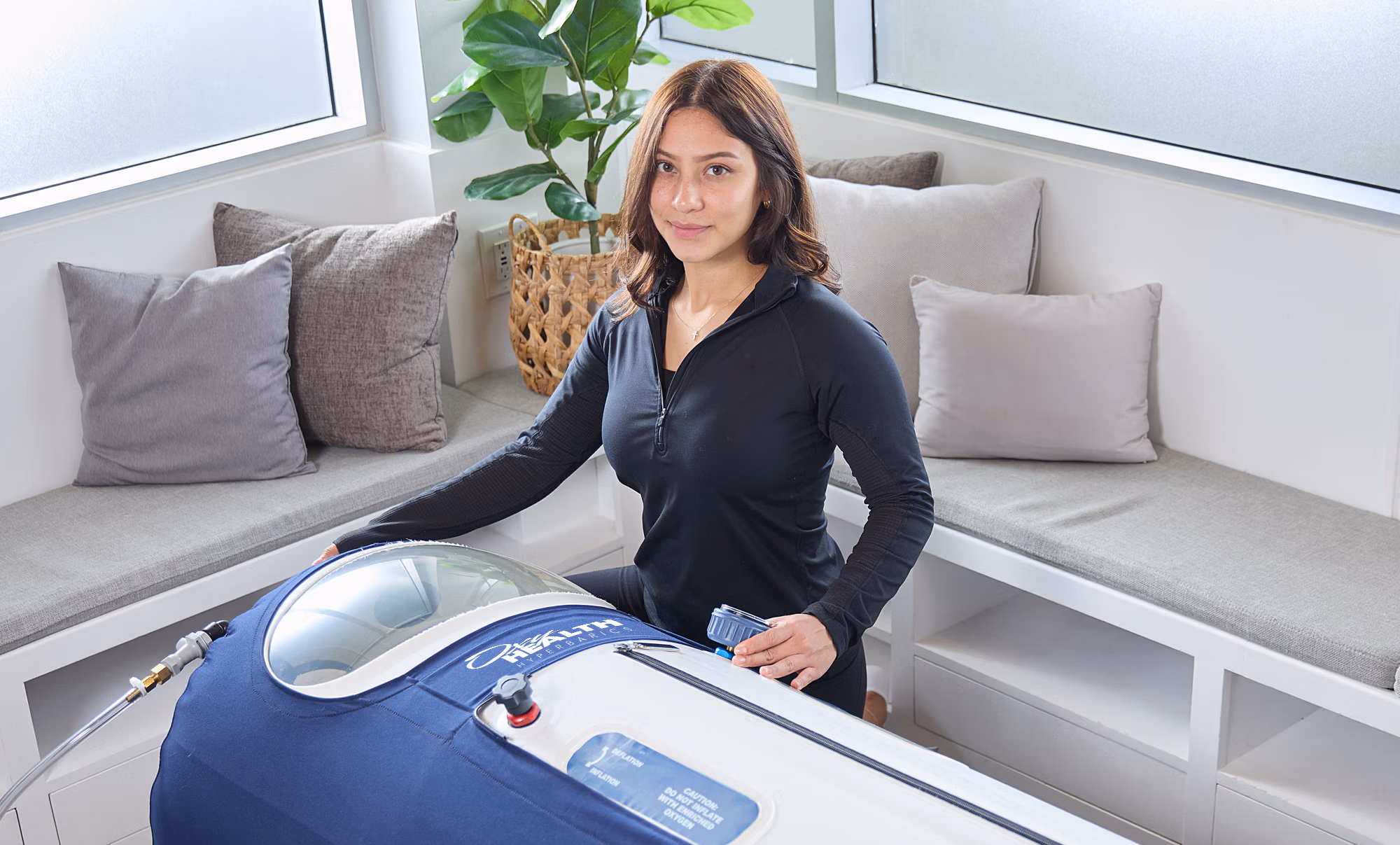 Young woman in black top sitting beside a blue and white hyperbaric oxygen therapy chamber in a bright, cushioned room.
