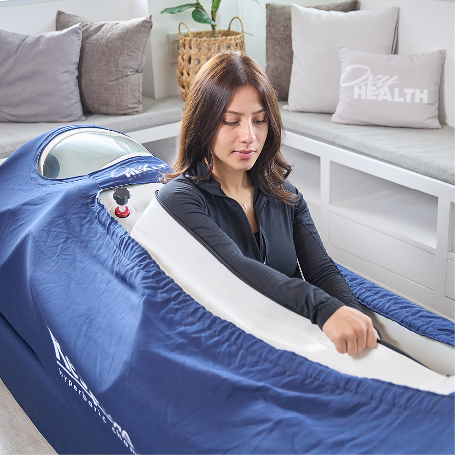 Woman using a blue hyperbaric chamber with a zippered cover in a bright room with gray cushions and a plant.