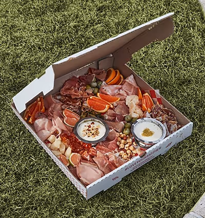 Open pizza box filled with charcuterie, cheeses, dips, and fruit