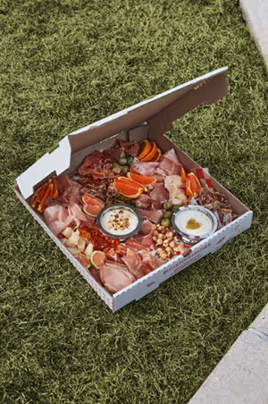 Open pizza box filled with charcuterie, cheeses, dips, and fruit