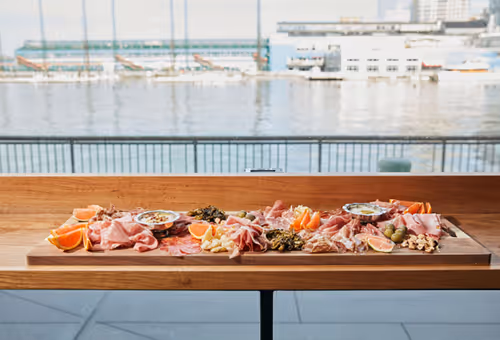 Long charcuterie board with meats, cheeses, and fruit set on a table by the water