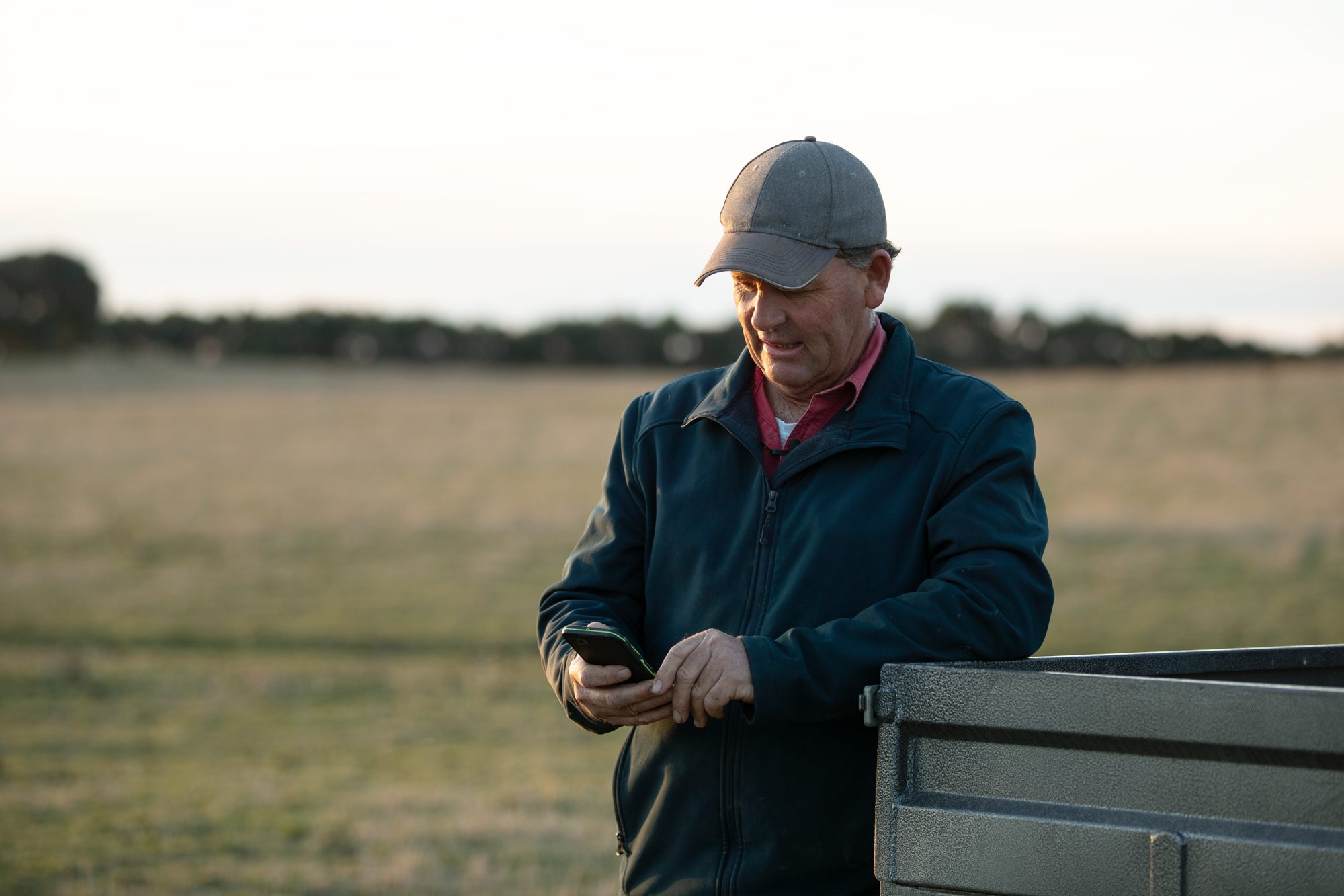 Farmer leaning on his ute in the paddock, checking his tank levels on his phone