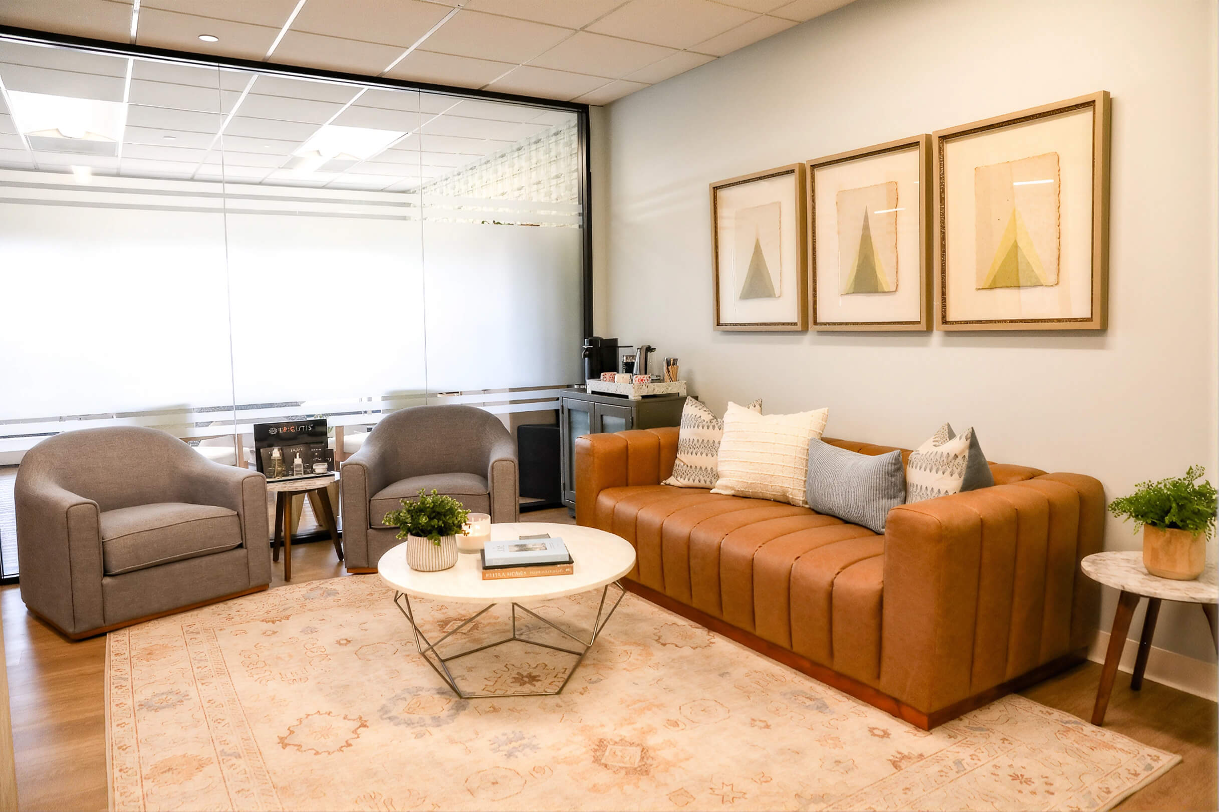 Bright medical or spa treatment room with white adjustable examination chair, rolling stool, wood flooring, three windows, and abstract artwork on walls.