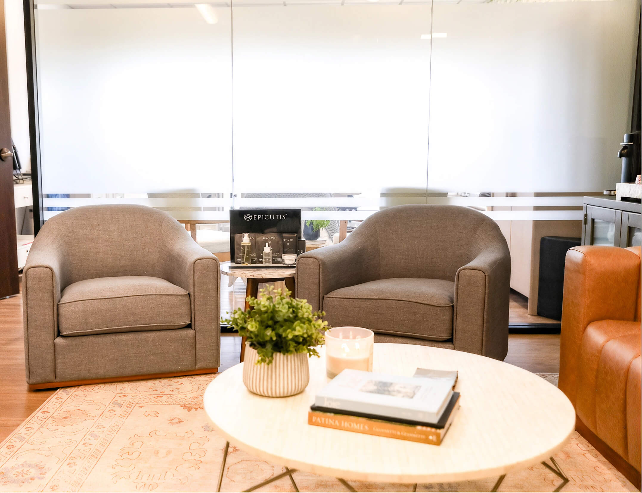 Modern waiting room with brown leather chairs, a round coffee table with a potted orchid, and a glass wall with a wooden door.