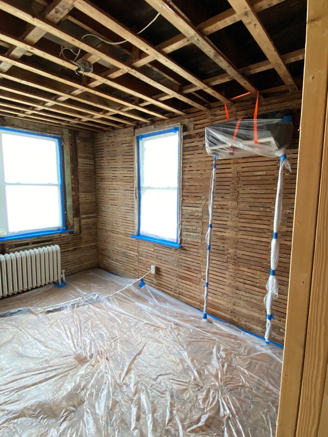 Room under renovation with stripped walls and ceiling, windows taped, and plastic covering floor and vent.