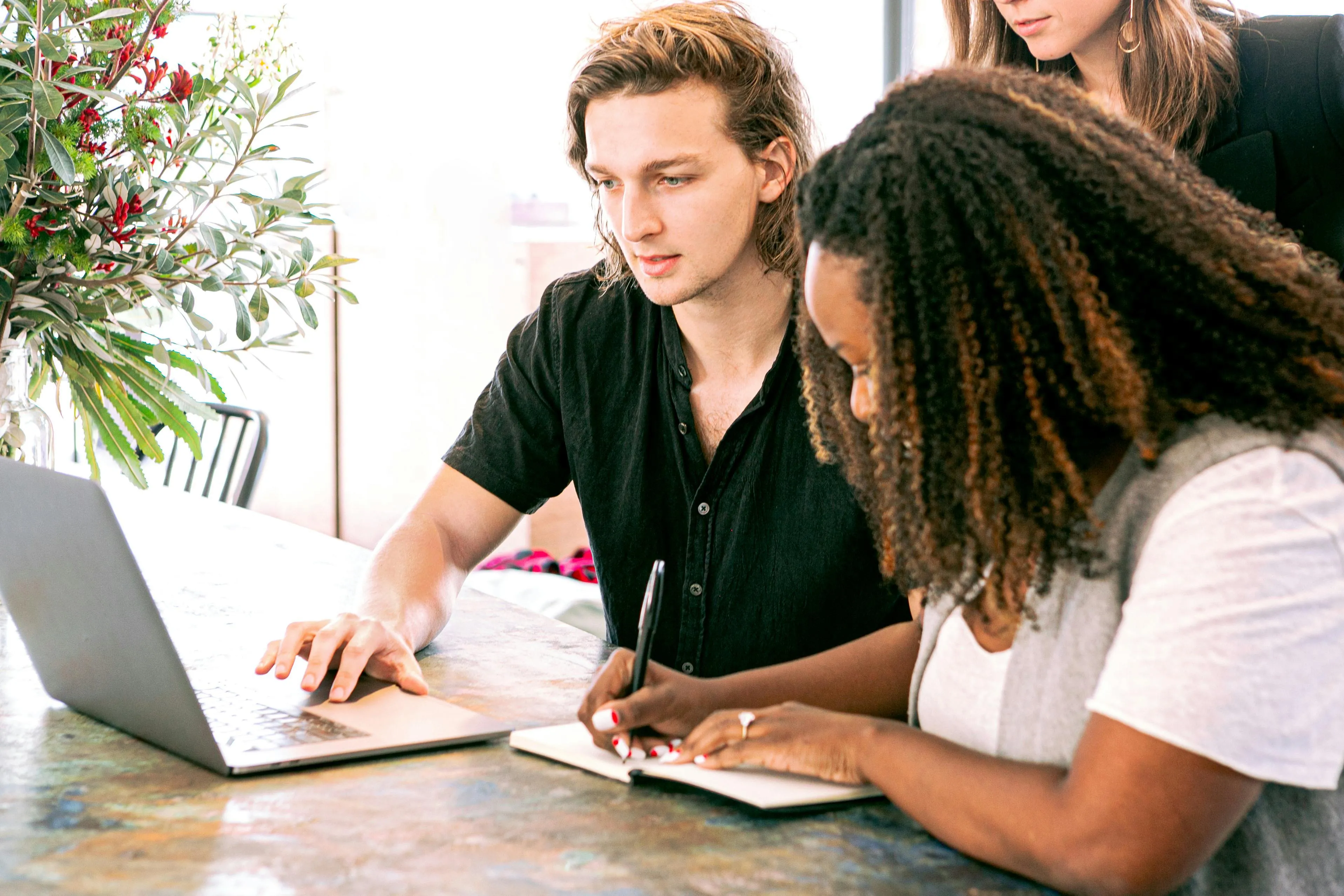 Three people working together at a table with a laptop and notebook, one person writing notes while others look on attentively.