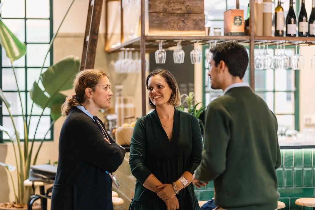 Three people engaged in a conversation inside a cozy, well-lit bar or cafe with green-tiled counters, hanging wine glasses, and bottles on shelves behind them.