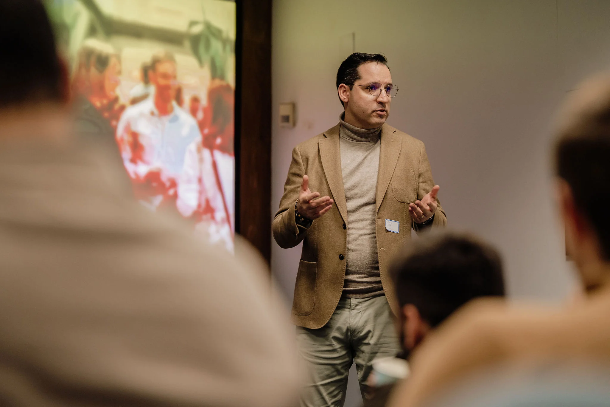 A man wearing glasses, a tan blazer, and a beige turtleneck sweater is speaking in front of an audience with a blurred projected image behind him.