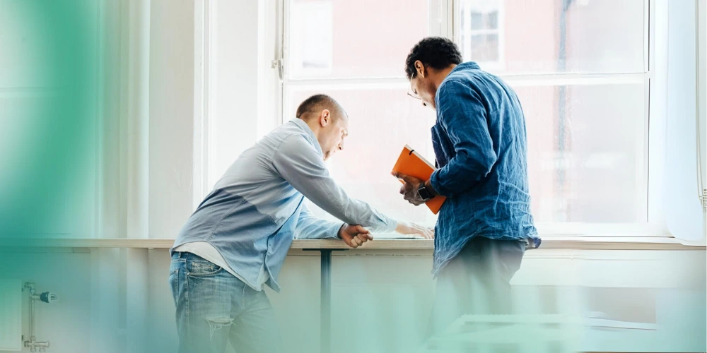 Two men in casual clothes engaged in discussion while standing by a window in a bright room, one holding an orange notebook.